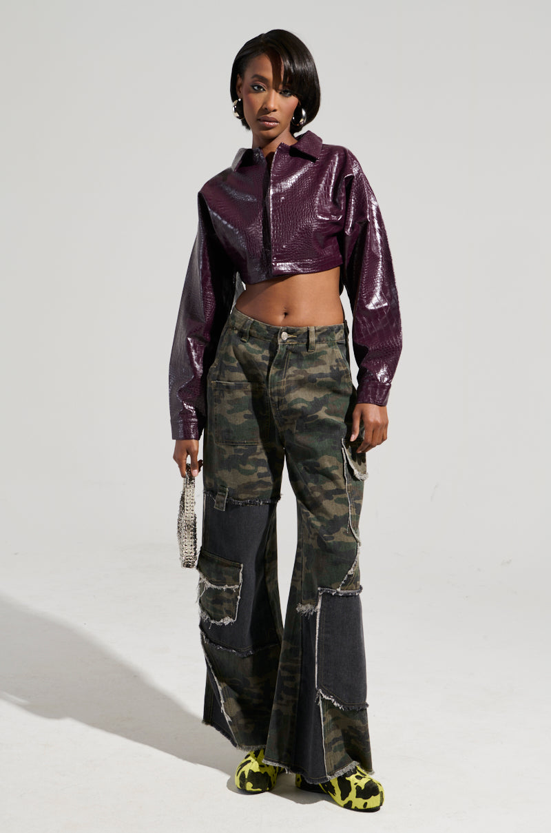 A person stands confidently against a plain background, wearing the sleek NATALIA CROCODILE PU BUTTON DOWN SHIRT and distressed camo pants. They hold a small bag and flaunt bright green and black shoes, exuding style with every step.