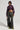 A person stands confidently against a plain background, wearing the sleek NATALIA CROCODILE PU BUTTON DOWN SHIRT and distressed camo pants. They hold a small bag and flaunt bright green and black shoes, exuding style with every step.