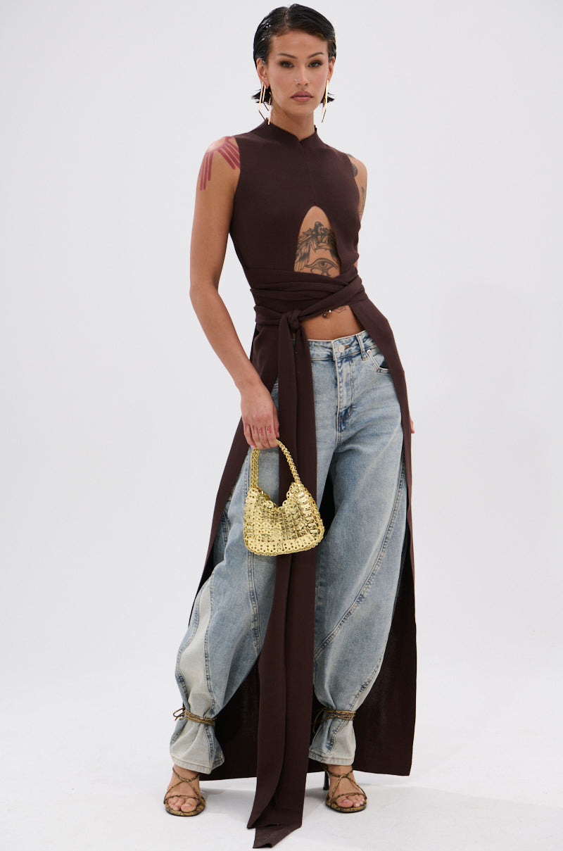 Wearing the OVER THE TOP CUTOUT SWEATER IN CHOCOLATE BROWN with a front slit and long sash, loose light blue jeans with tied cuffs, heeled sandals, and holding a small gold beaded handbag, the person poses against a plain white background. 0