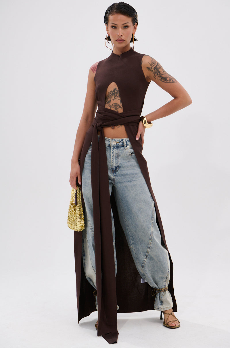 A person exudes confidence in the OVER THE TOP CUTOUT SWEATER IN CHOCOLATE BROWN, paired with light blue loose-fit jeans, strappy heels, and a gold handbag. They flaunt tattoos, slicked-back hair, hoop earrings, and a bold look. 1
