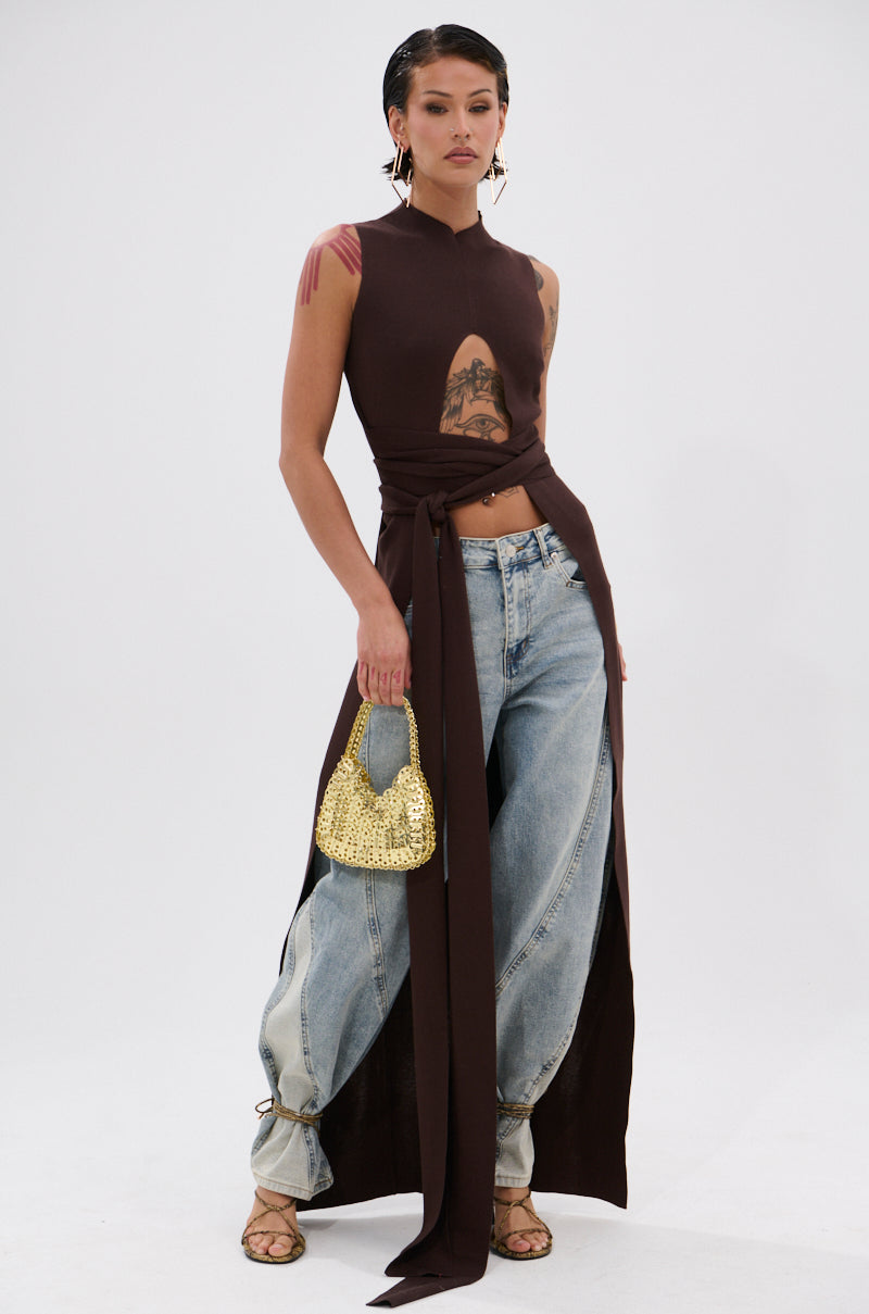 A woman wears the OVER THE TOP CUTOUT SWEATER IN CHOCOLATE BROWN with a front cutout and long draping ties, paired with light blue baggy jeans tied at the ankles, heeled sandals, gold hoop earrings, and a small gold handbag. 2