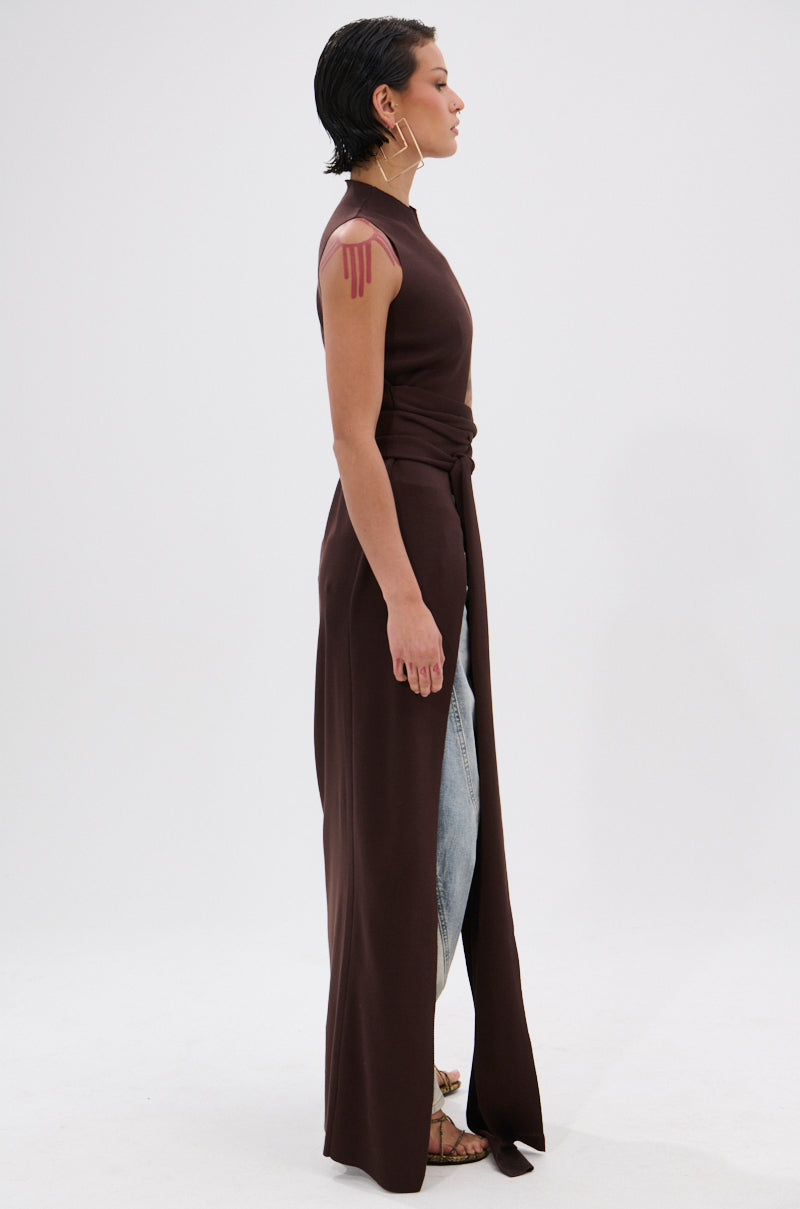 A person with short dark hair stands in profile against a white background, wearing the OVER THE TOP CUTOUT SWEATER IN CHOCOLATE BROWN over light blue jeans and sandals, with visible pink designs on their upper arm. 3