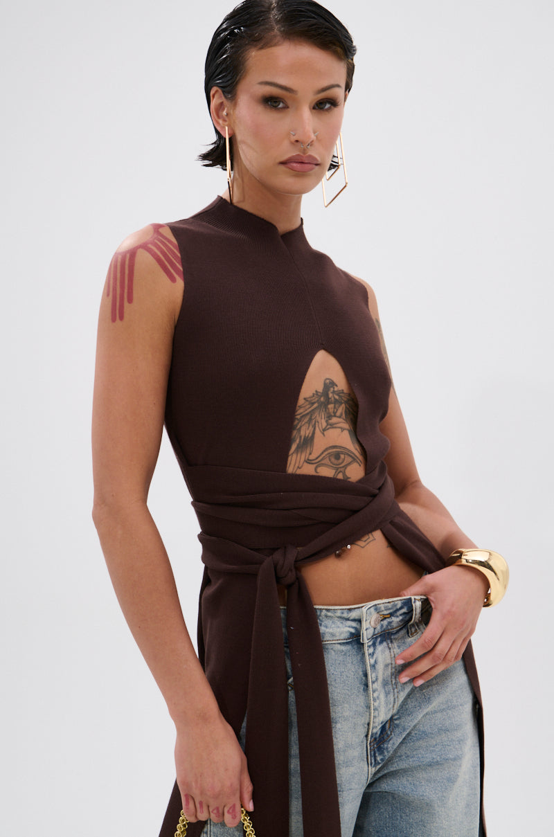 A woman with short dark hair and visible tattoos wears the OVER THE TOP CUTOUT SWEATER IN CHOCOLATE BROWN, paired with light blue jeans, geometric earrings, and a gold bracelet. 6