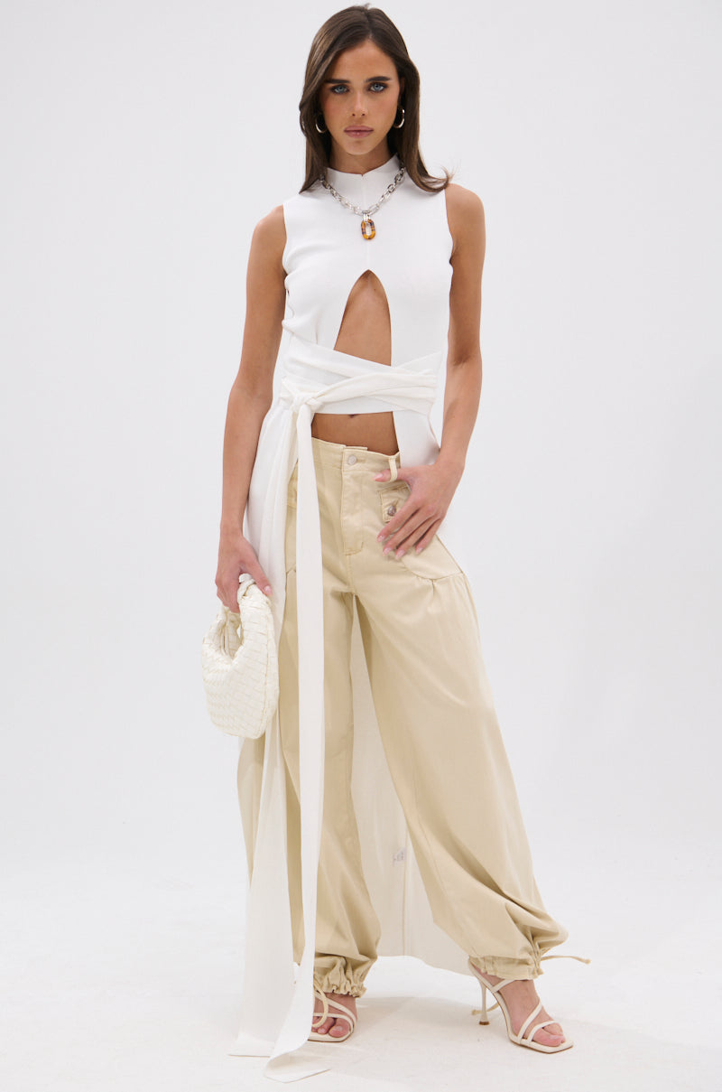 A woman poses in the OVER THE TOP CUTOUT SWEATER—sleeveless, white, with long ties—paired with wide-leg beige pants with gathered hems, white heeled sandals, and a small white woven handbag against a plain background. 0