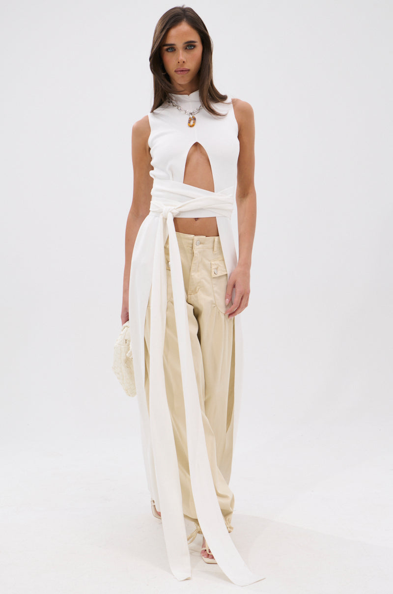 A woman stands against a white background wearing the OVER THE TOP CUTOUT SWEATER—a sleeveless white top with a high neckline and front cutout—paired with beige wide-leg trousers, white sandals, and holding a textured handbag. 1