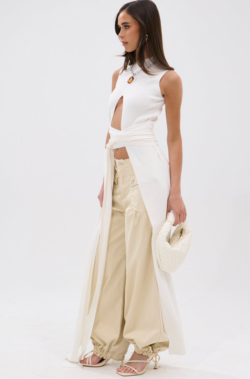 A woman wears the OVER THE TOP CUTOUT SWEATER in white, featuring a ribbed knit, sleeveless style, front cutout, and long draped panels. She pairs it with beige ankle-cinched wide-leg pants, heeled sandals, a woven white clutch, and minimal jewelry. 2