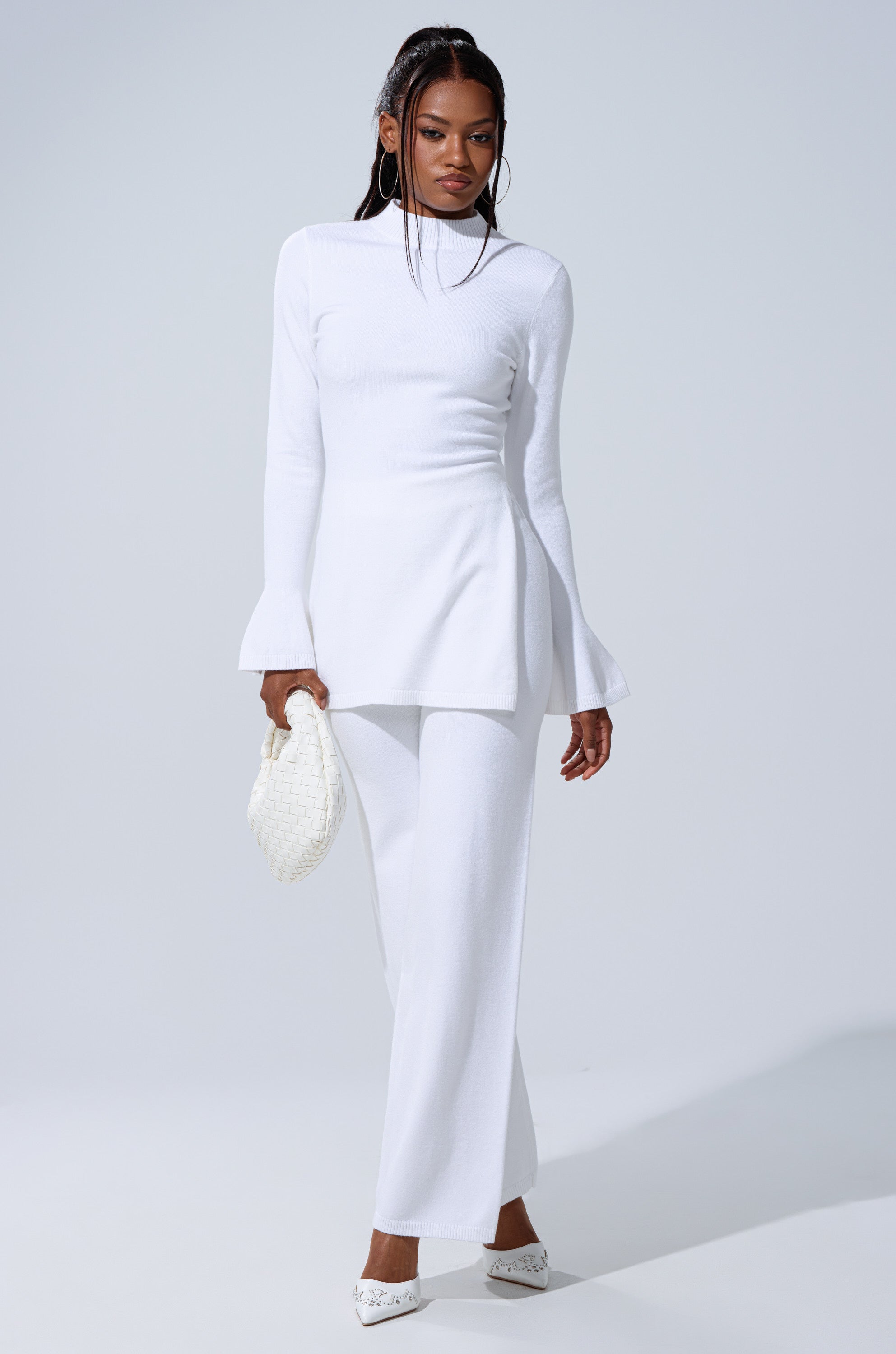 Against a light gray backdrop, a woman models the GET COZY WITH ME WIDE LEG PANT IN WHITE with a fitted white tunic, flared cuffs, white shoes, and a textured clutch. 0