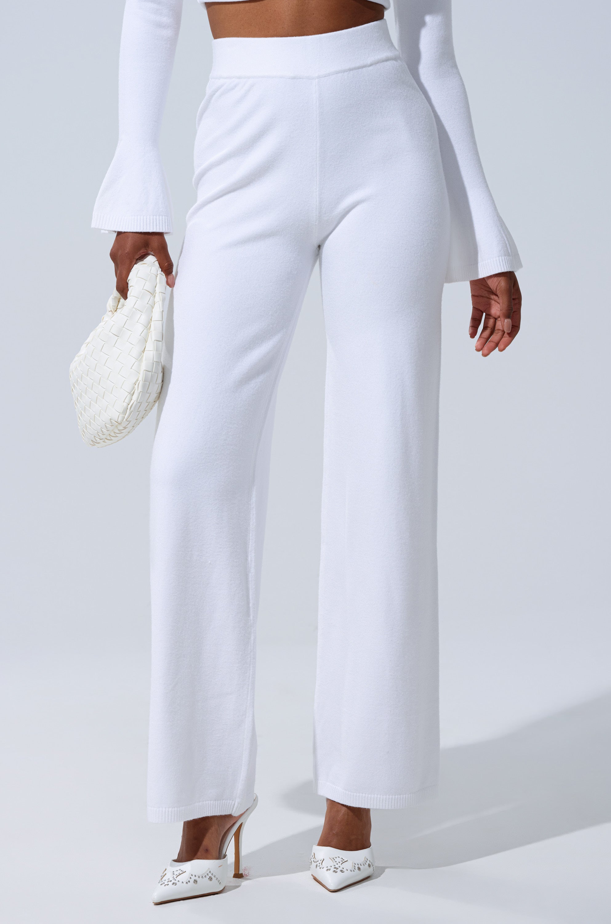 A person wears the GET COZY WITH ME WIDE LEG PANT IN WHITE with a long-sleeve white top, flared cuffs, white pointed-toe heels, and holds a woven white clutch. The background is plain and light. 1