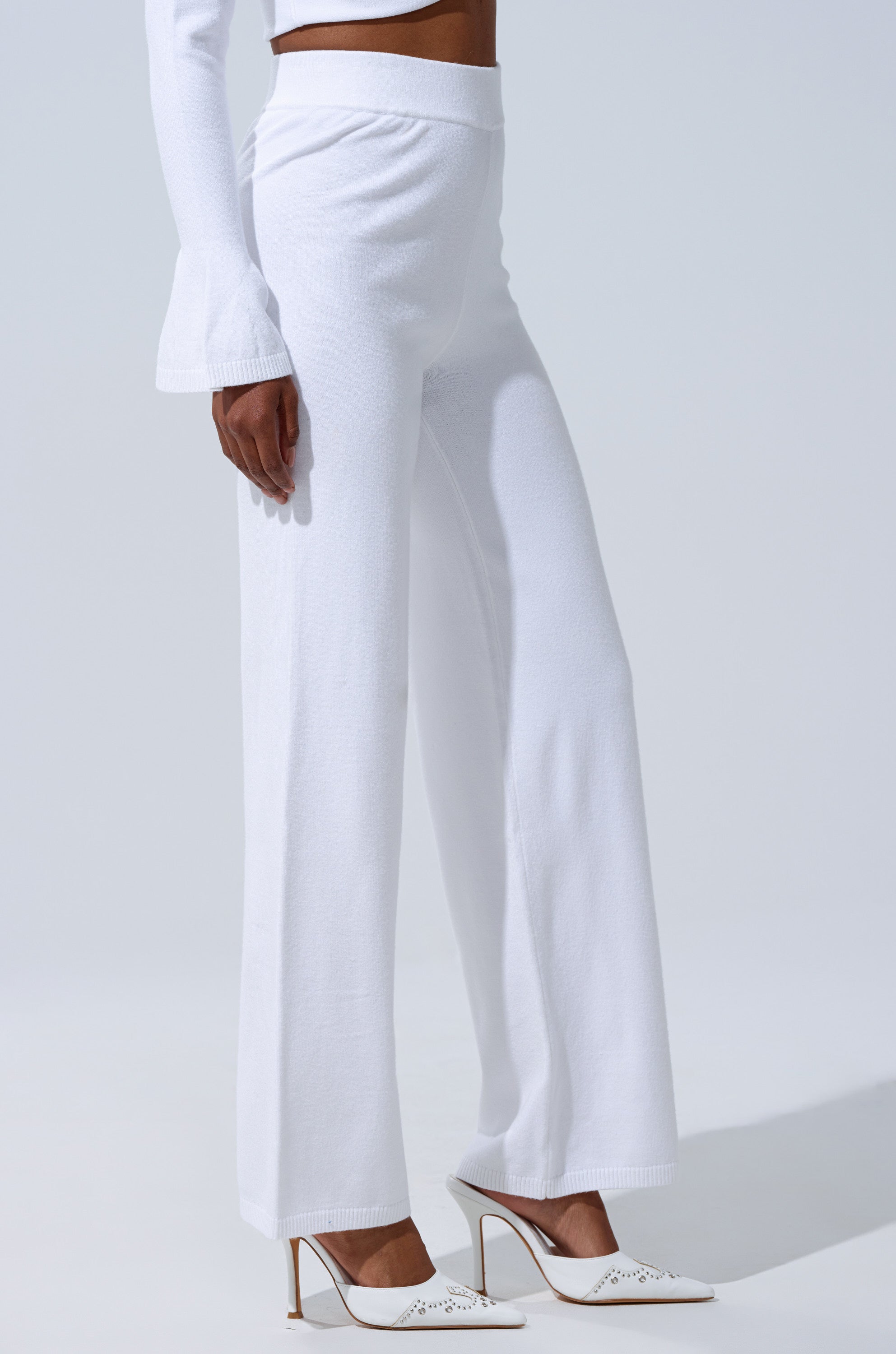 A person wears the GET COZY WITH ME WIDE LEG PANT IN WHITE, paired with a long-sleeve white top with flared cuffs and white pointed-toe heels, standing against a plain light background. 2