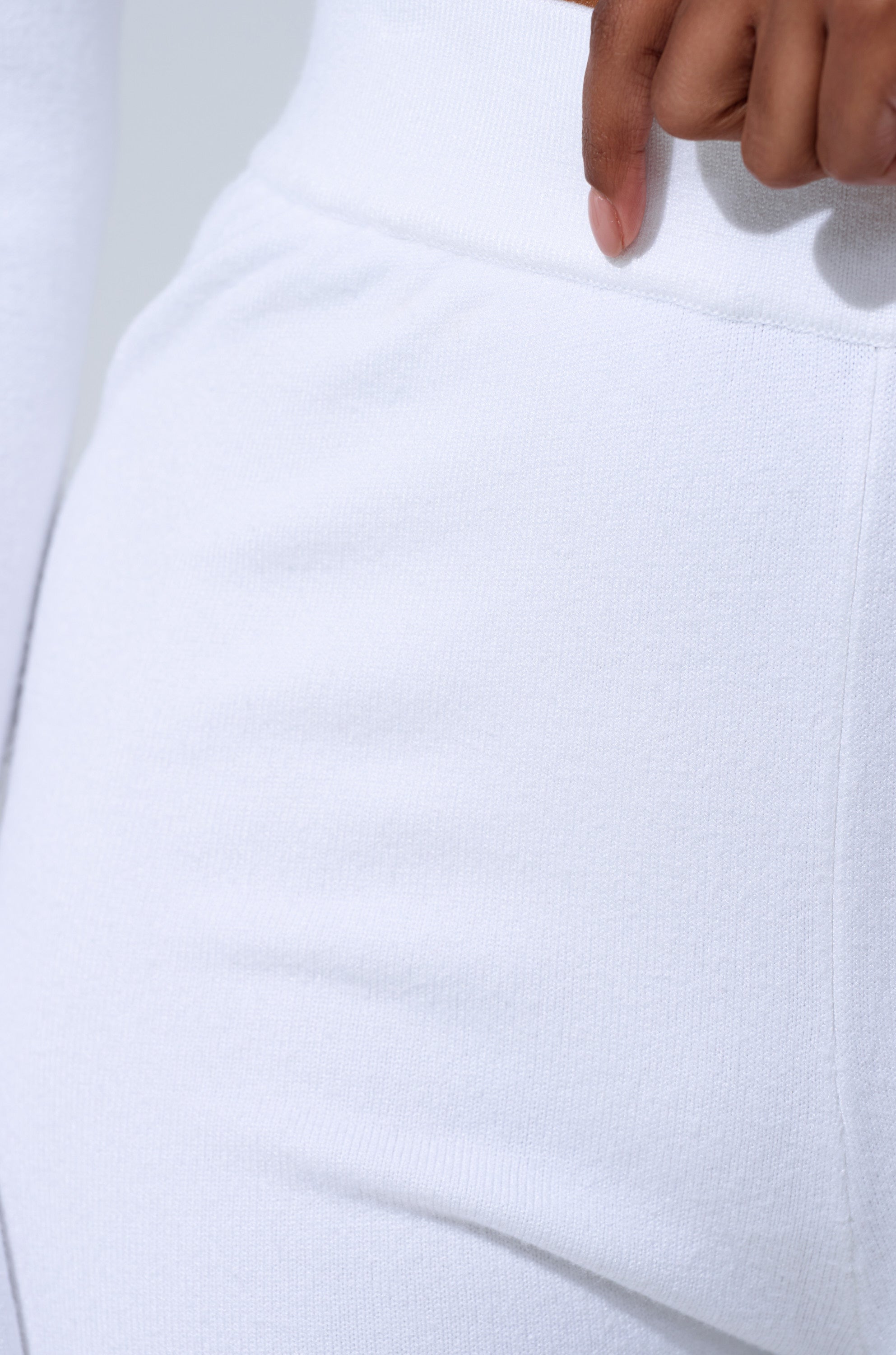 A close-up of a person wearing the GET COZY WITH ME WIDE LEG PANT IN WHITE—fitted, high-rise, with a matching white top. Their hand rests gently at the waist against a plain, light background. 5