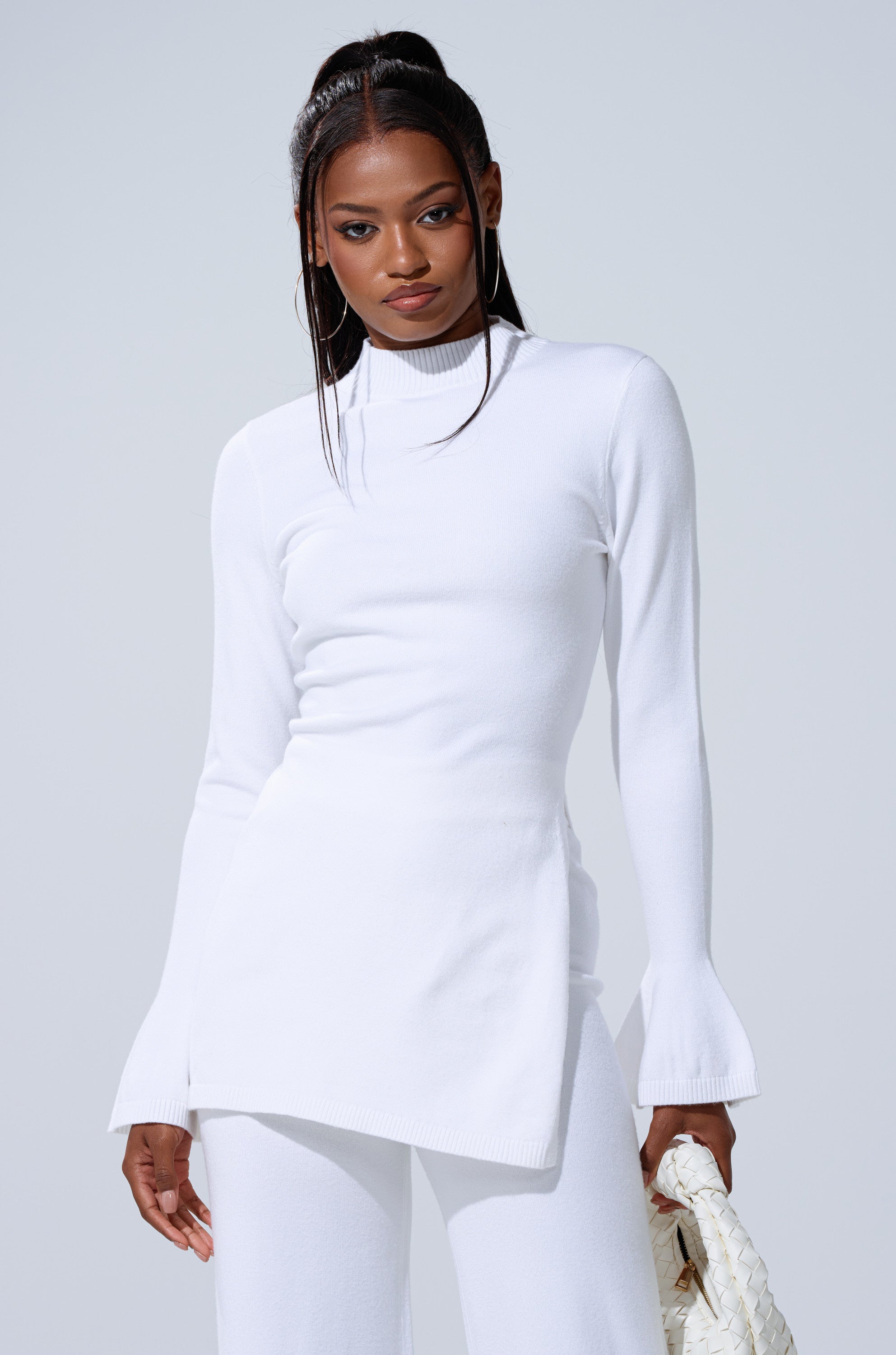 A woman with long dark hair in a high ponytail wears the GET COZY WITH ME OVERSIZED SWEATER IN WHITE with matching white pants, holding a textured white handbag against a plain light background. 0