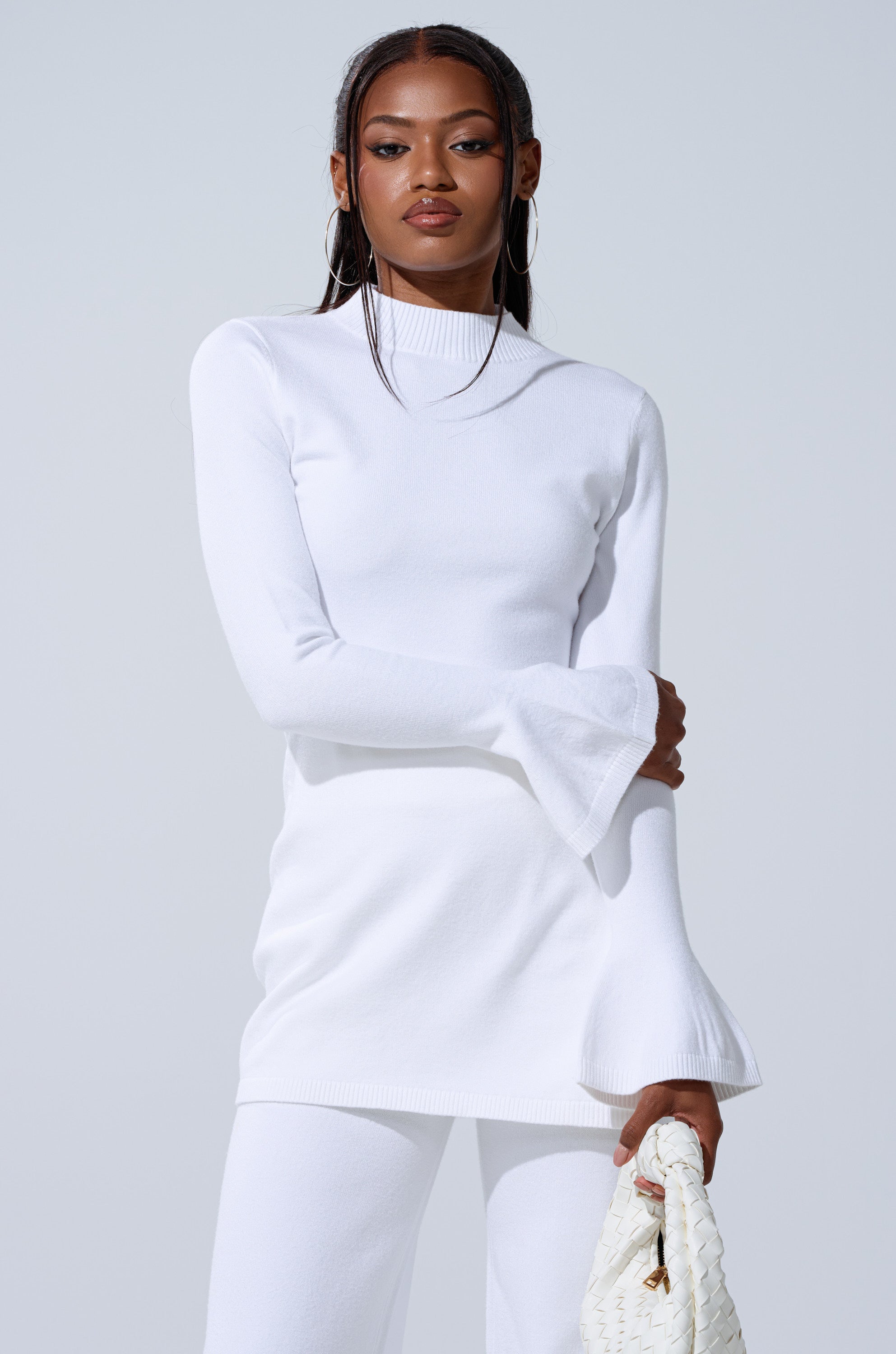 A woman stands against a light gray background, wearing the GET COZY WITH ME OVERSIZED SWEATER IN WHITE and matching pants, holding a white woven handbag and looking confidently at the camera. 1