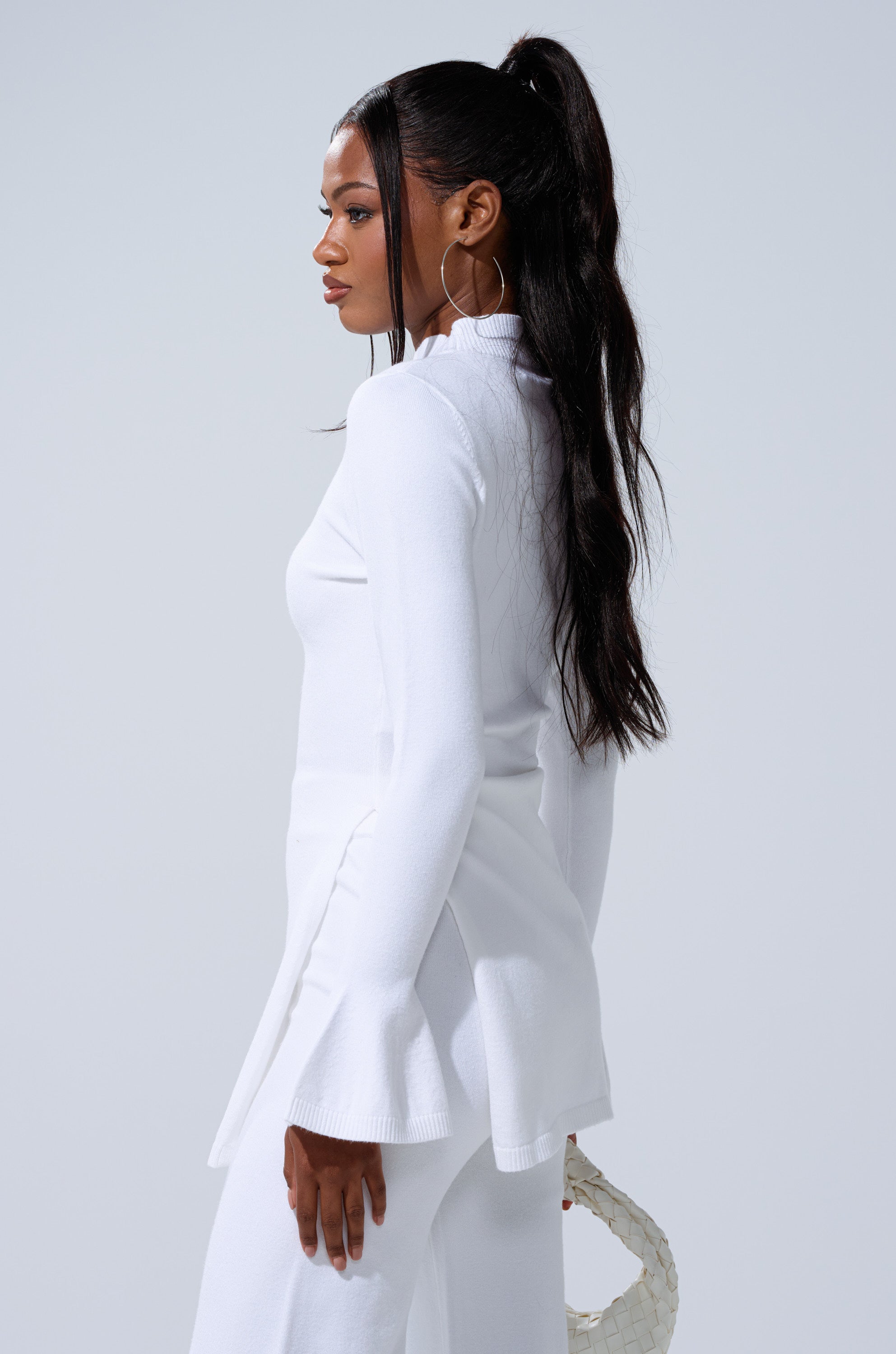 A woman with long, dark hair in a high ponytail wears the GET COZY WITH ME OVERSIZED SWEATER IN WHITE and matching pants, holding a textured handbag for a chic loungewear look against a plain light gray background. 2