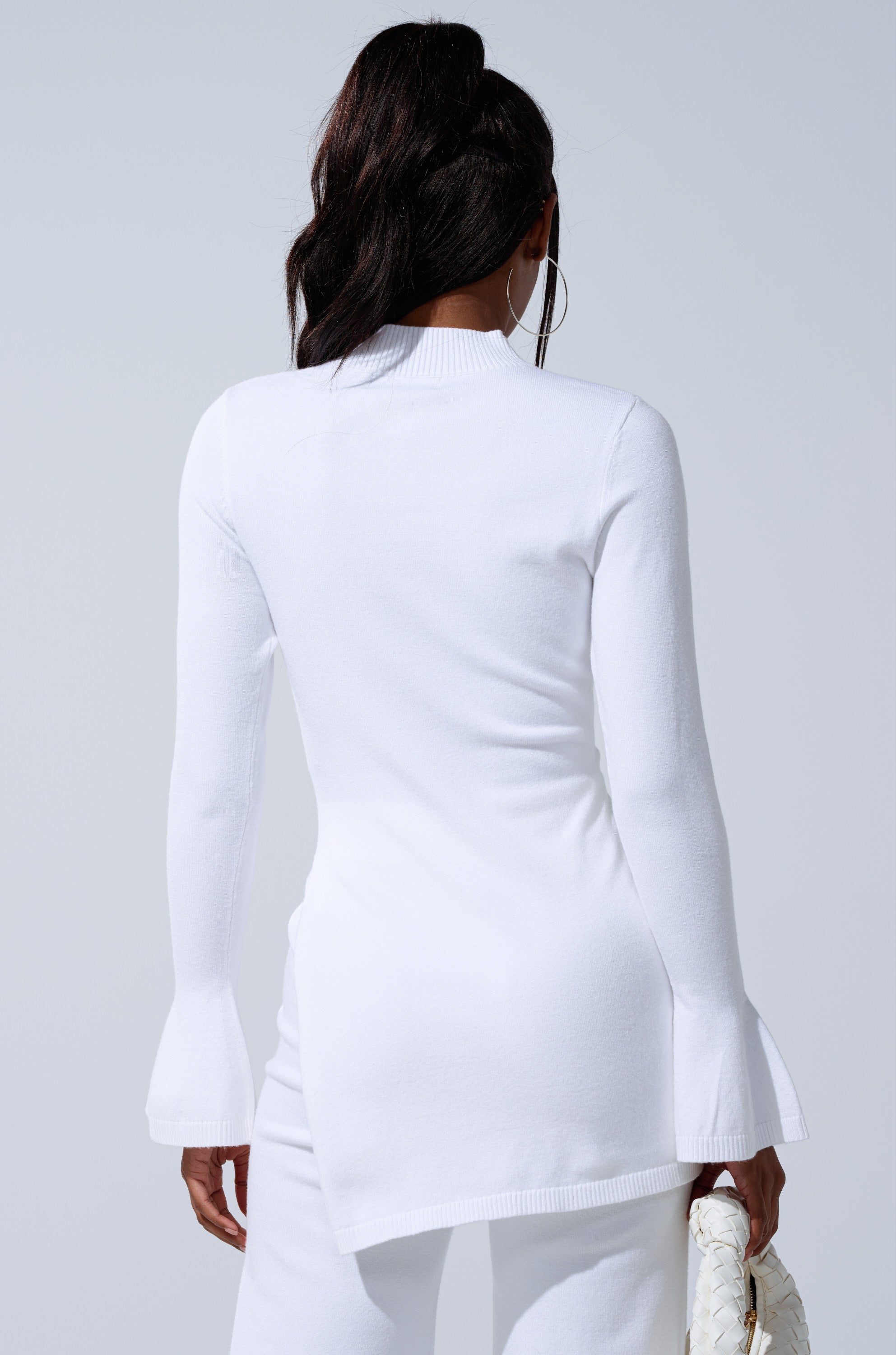 A woman is seen from the back in the GET COZY WITH ME OVERSIZED SWEATER IN WHITE and matching pants, carrying a white woven handbag. Her dark hair is styled in a high ponytail, highlighting her relaxed loungewear look. 3
