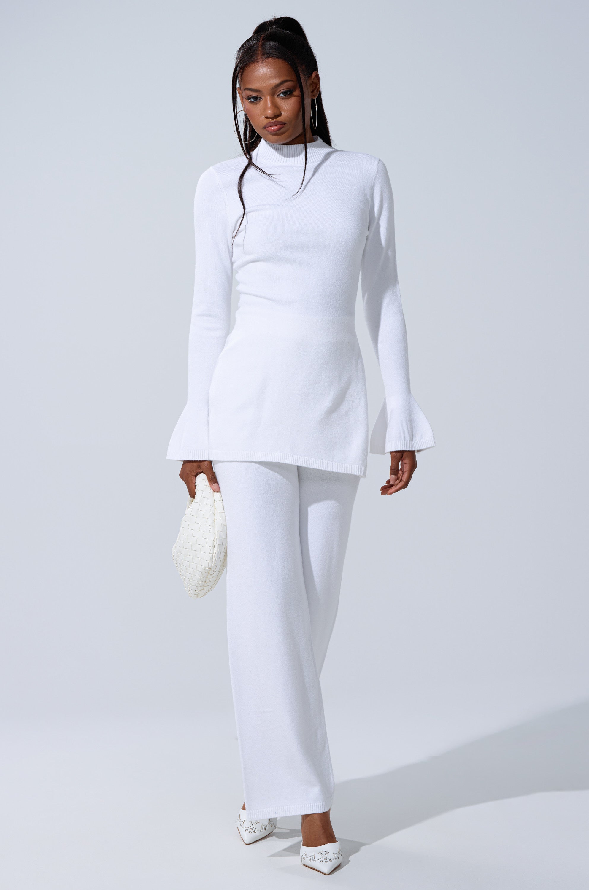 A woman wears the GET COZY WITH ME OVERSIZED SWEATER IN WHITE, paired with white wide-leg pants and shoes. She holds a textured clutch, exuding effortless loungewear elegance against a light background. 5