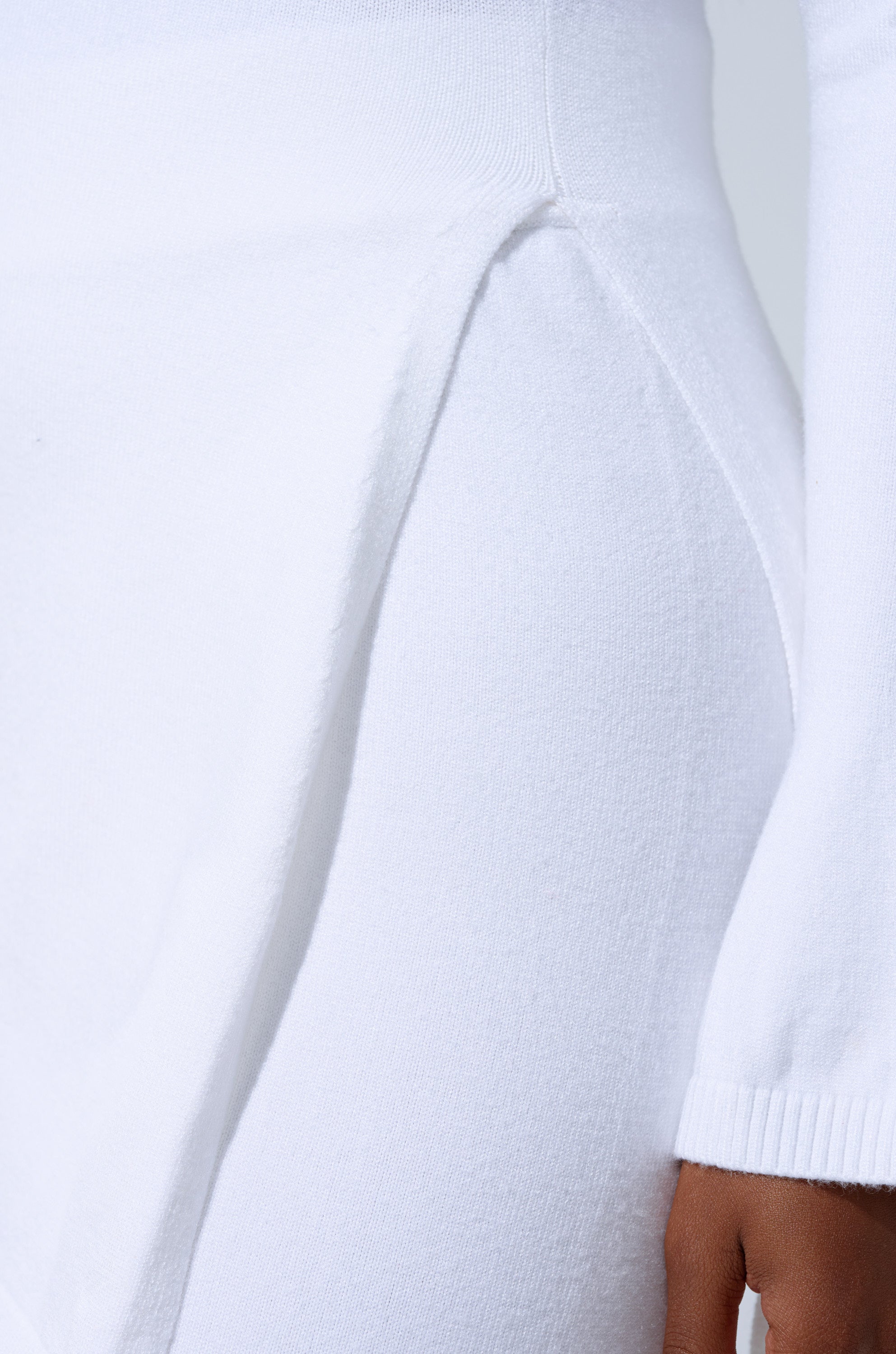 Close-up of a person wearing the GET COZY WITH ME OVERSIZED SWEATER IN WHITE, featuring textured draped detail at the waist and long sleeves. The background is plain and light-colored. 6