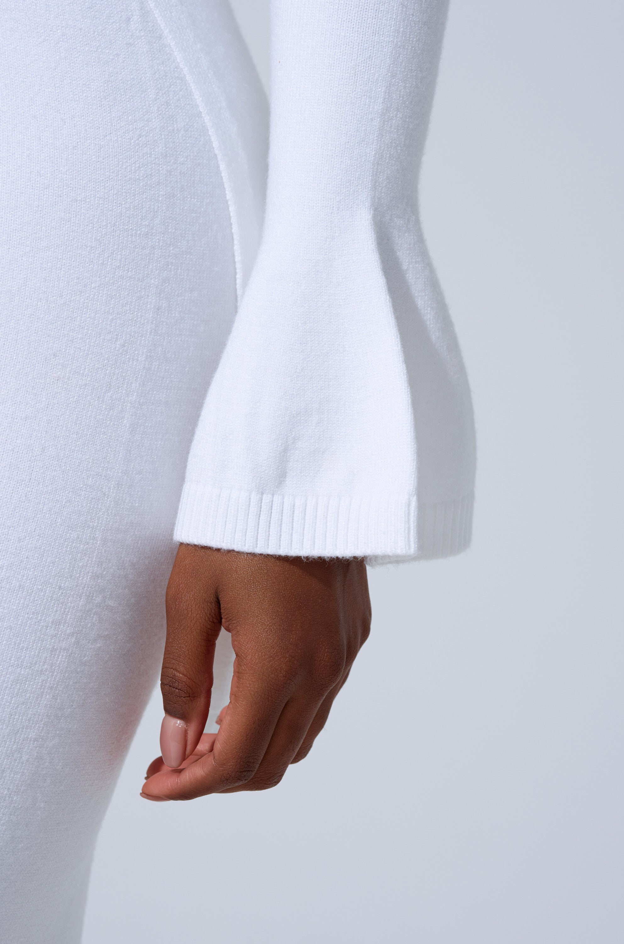 A close-up showcases the GET COZY WITH ME OVERSIZED SWEATER IN WHITE, featuring a relaxed fit and flared ribbed sleeves. The wearer stands against a light gray background with their hand resting gently by their side. 7