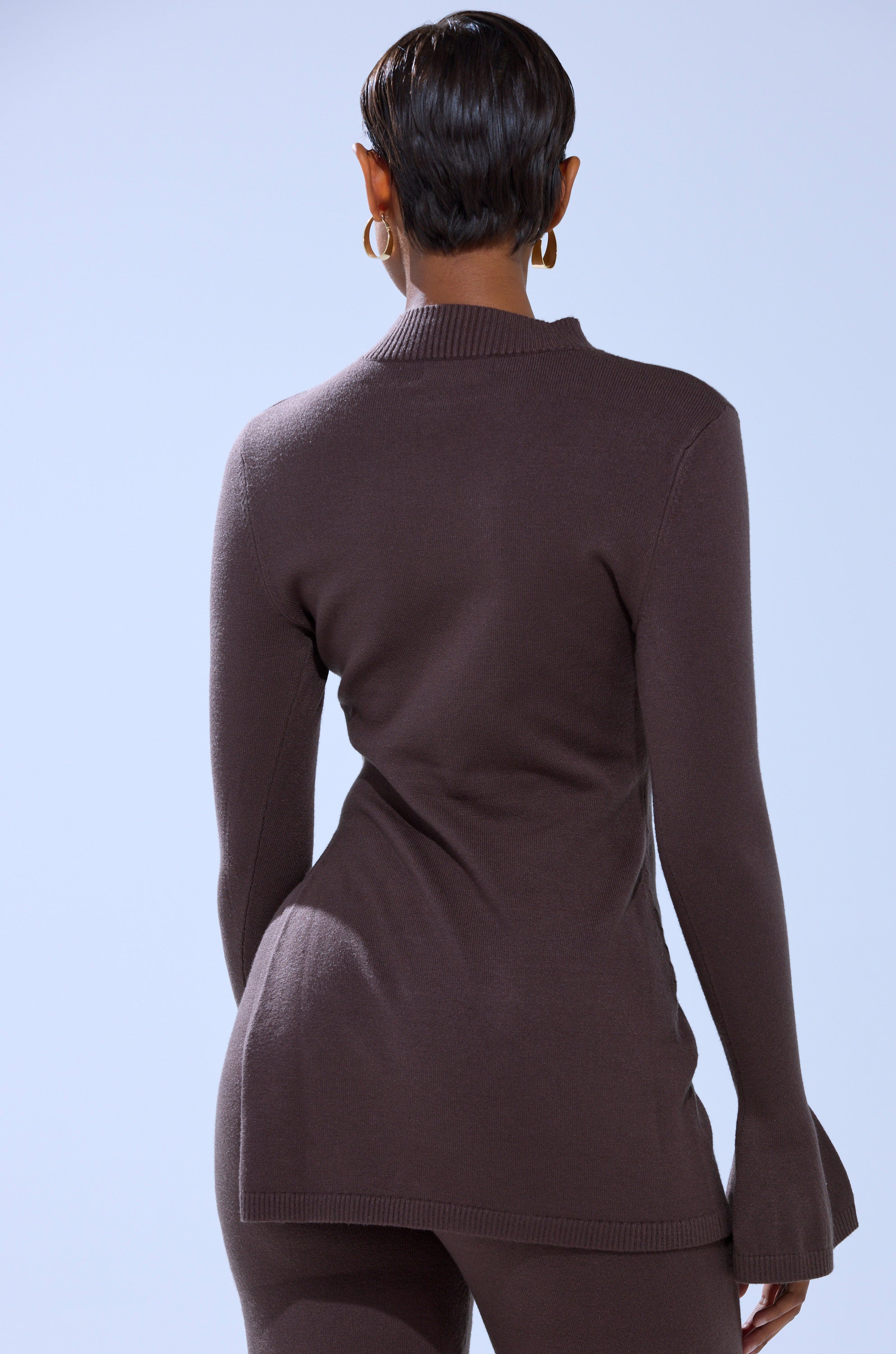 A person with short hair and gold hoop earrings wears the AKIRA Label GET COZY WITH ME OVERSIZED SWEATER and matching pants, standing with their back to the camera against a light blue background. 5