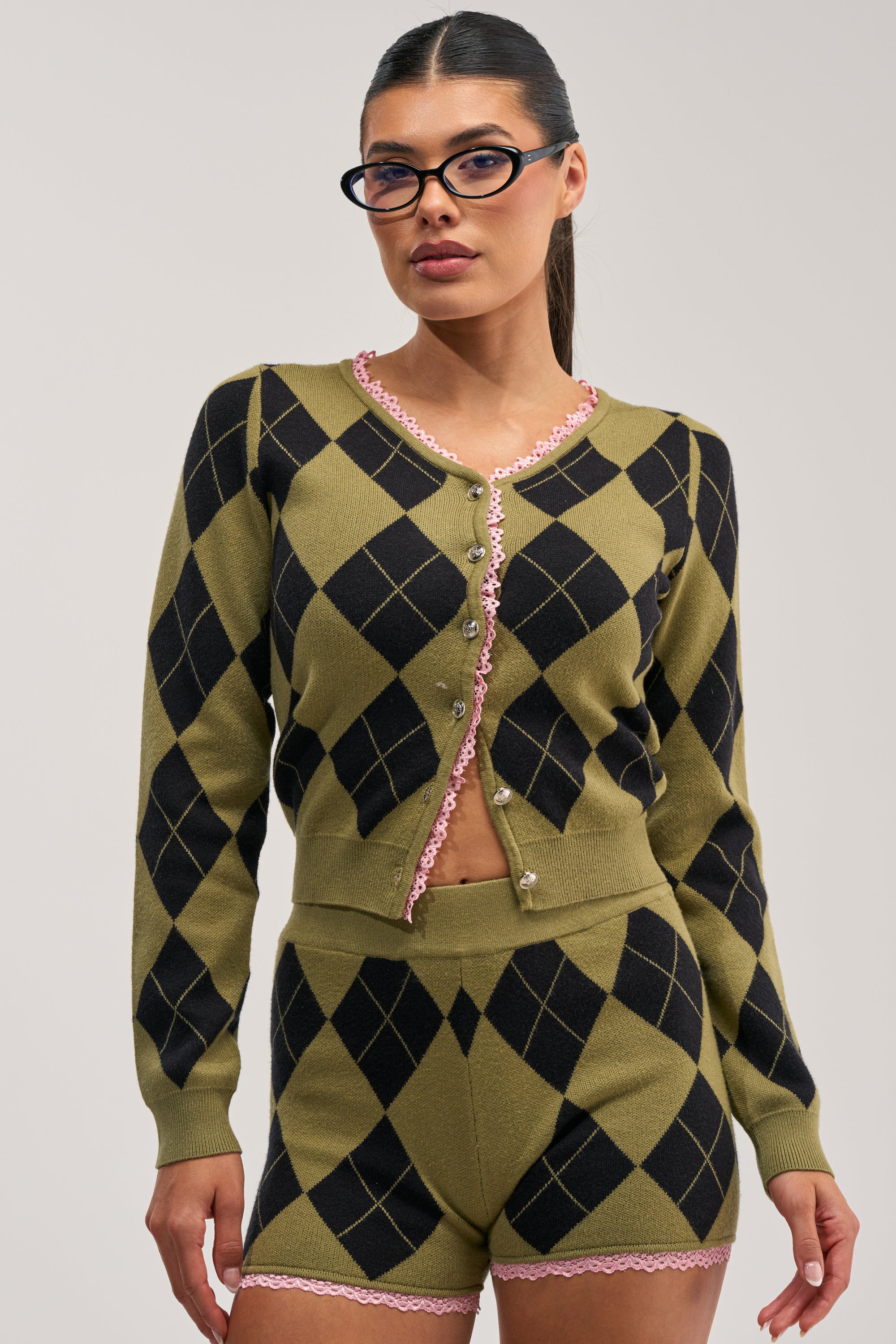 Woman with glasses wearing the CRAZY FOR YOU ARGYLE CARDIGAN—a preppy-chic green knit cardigan with matching high-waisted shorts featuring pink trim—standing against a plain light background.