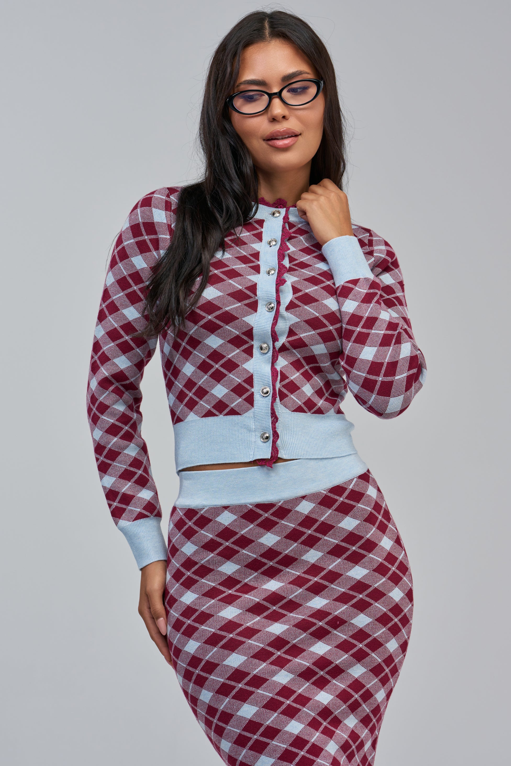 A woman wearing glasses and the AVA ARGYLE CARDIGAN—a purple and white argyle cropped knit cardigan with light blue trim—along with a matching skirt, stands against a plain gray background.
