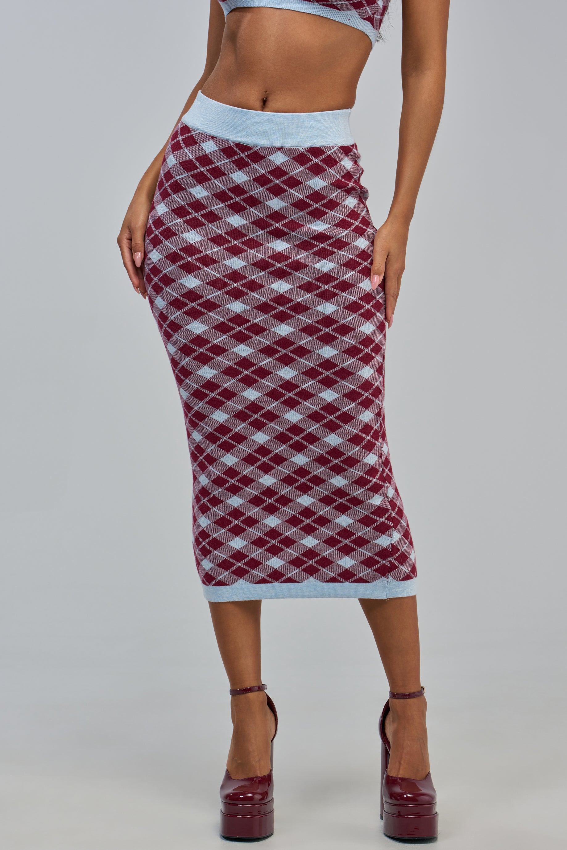 A woman is seen wearing the AVA ARGYLE MAXI SKIRT—a high-waisted, ankle-length skirt showcasing a striking combination of burgundy and light blue in a classic argyle pattern. Paired with a matching crop top and burgundy platform heels, she stands against a plain background, highlighting the preppy-chic style and modern appeal of this standout piece.