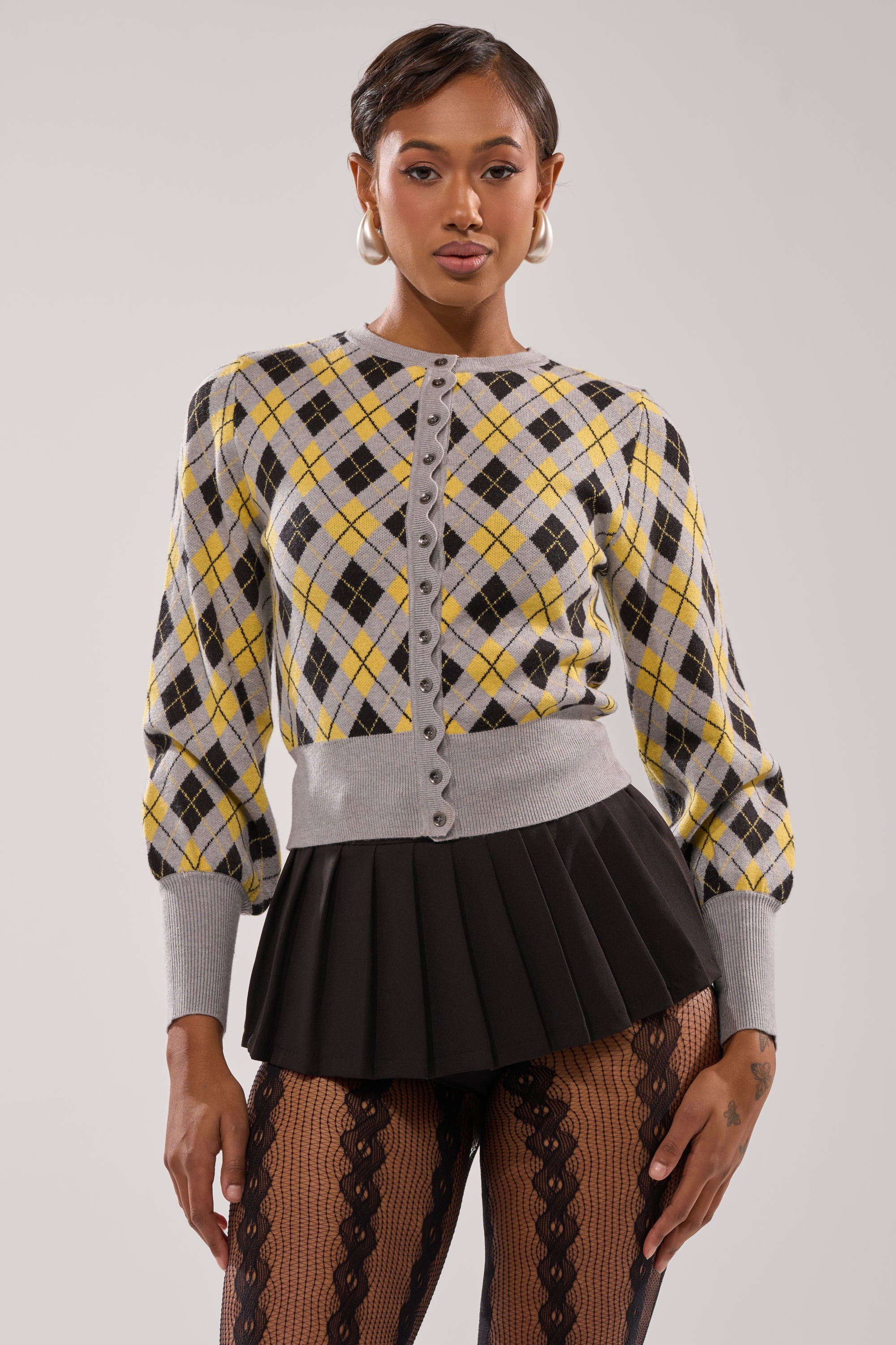 A woman wearing the GUESS WHAT PLAID CARDIGAN in yellow, gray, and black with gray trim, a black pleated mini skirt, and black lace tights stands against a light background, confidently facing the camera. 0