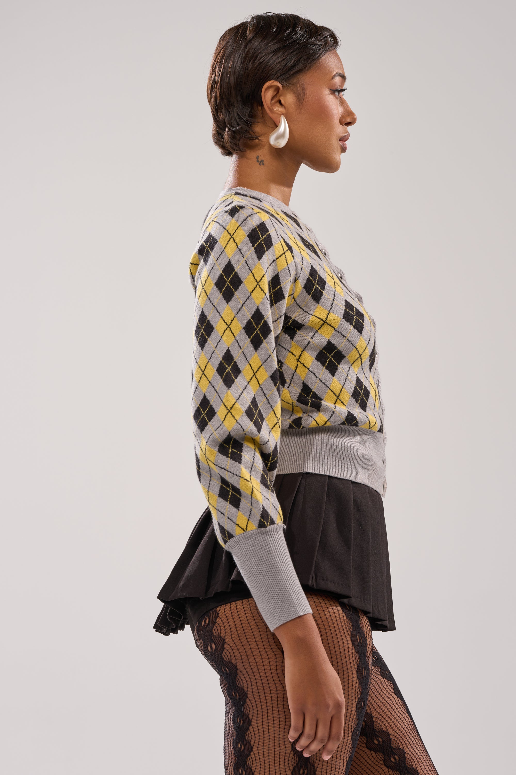 A woman in profile wears the GUESS WHAT PLAID CARDIGAN in gray and yellow, paired with a black ruffled skirt and patterned tights. Her short hair, white hoop earrings, and neck tattoo complete a preppy look against a plain light background. 2