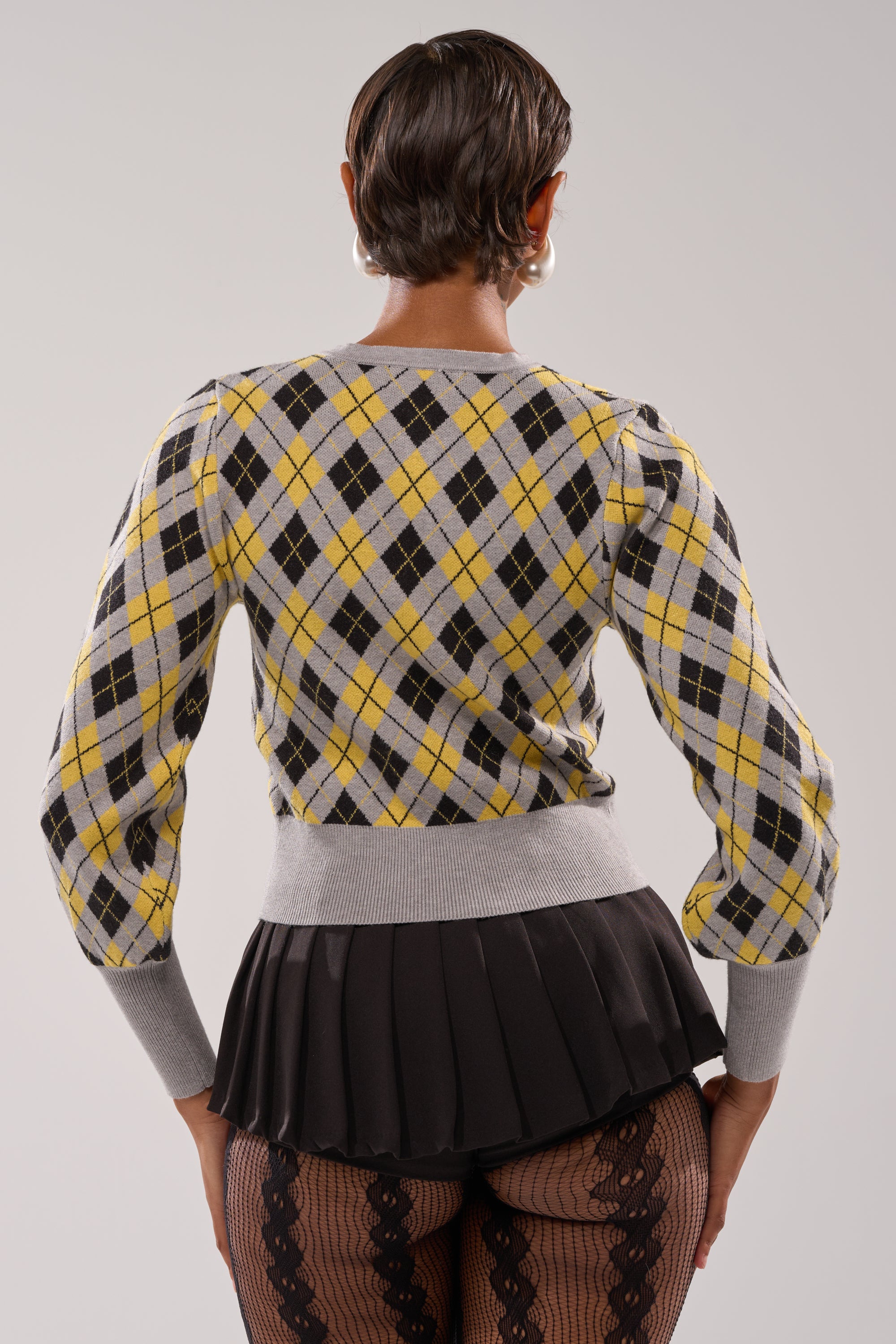 A person with short hair is seen from the back, wearing the GUESS WHAT PLAID CARDIGAN in yellow and gray, a black pleated mini skirt, and patterned black tights against a plain light background. 3