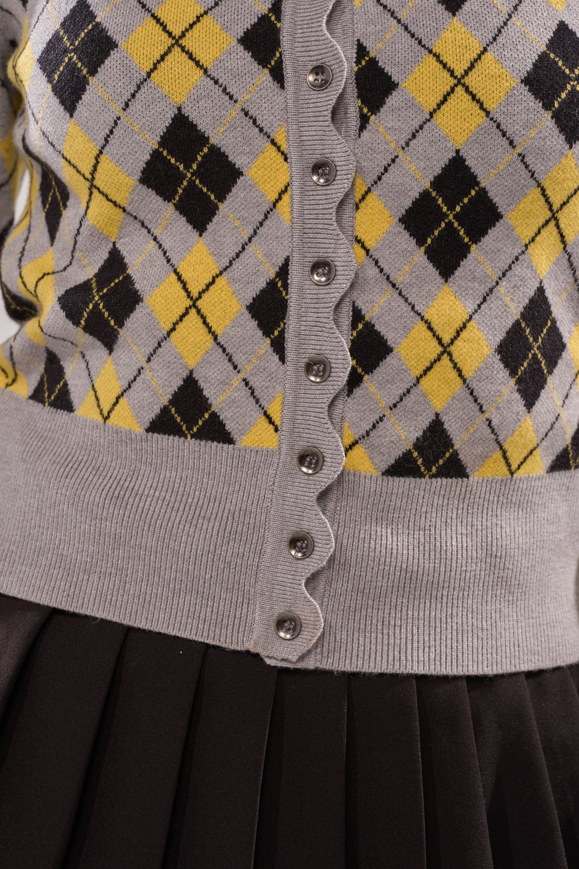 Close-up of the GUESS WHAT PLAID CARDIGAN featuring yellow, black, and gray argyle pattern, decorative silver buttons, and ribbed waistband for a preppy look, styled with a black pleated skirt. 6