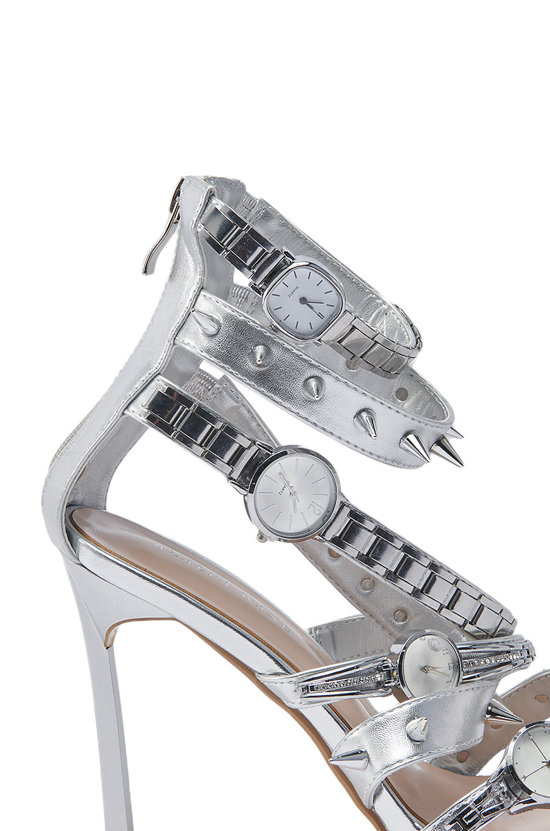 The AZALEA WANG ROBINSON SILVER WATCH SHOE STILETTO SANDAL features metallic silver faux leather straps, each accented with silver wristwatches and spikes. White watch faces with silver details give this sandal a bold, edgy look.