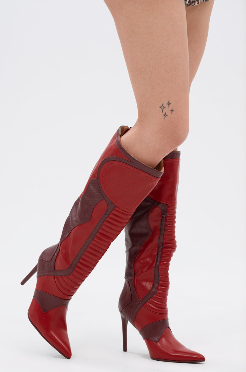 A fashion-forward individual strikes a confident pose in the AZALEA WANG RODRIGO RED BOOT STILETTO, featuring textured and smooth knee-high panels. Three black star tattoos accent their lower leg against a plain white background.