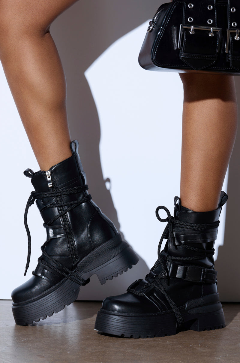 A person wearing the AZALEA WANG ROISIN BLACK COMBAT BOOTIE with chunky laces and side zippers stands on a wooden floor against a white background. Nearby, a black handbag with silver buckles complements the edgy style of the vegan leather booties.
