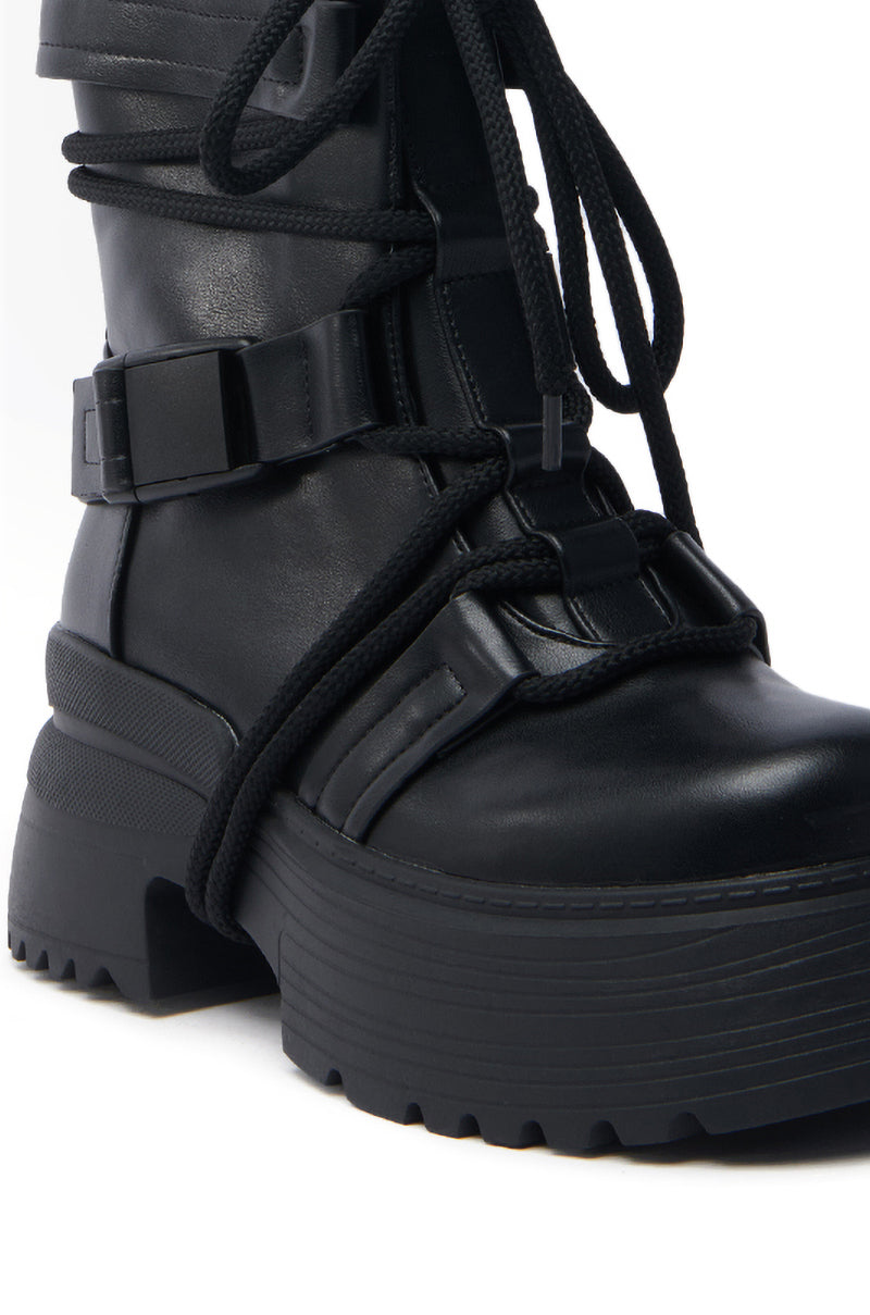 The AZALEA WANG ROISIN BLACK COMBAT BOOTIE is a black vegan leather bootie with thick laces and a chunky heel, featuring an ankle strap with buckle details for a rugged look. It has a textured, ridged sole designed for grip.