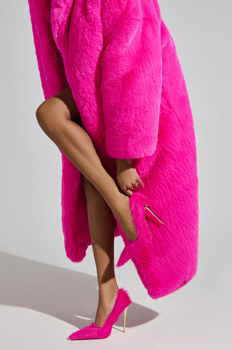A stylish individual dons a vibrant pink fur coat with AZALEA WANG ROMEE FUCHSIA PONY HAIR PUMPS, exuding elegance. They lift a foot to adjust the pointed toe of their stiletto heel, standing against a neutral gray background.