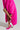A stylish individual dons a vibrant pink fur coat with AZALEA WANG ROMEE FUCHSIA PONY HAIR PUMPS, exuding elegance. They lift a foot to adjust the pointed toe of their stiletto heel, standing against a neutral gray background.