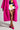 A person stands against a neutral background, wearing a vibrant pink faux fur coat, a colorful geometric-patterned mini skirt, and AZALEA WANG ROMEE fuchsia pony hair pumps with pointed toes. Only the lower half of the body is visible.