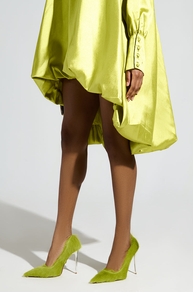 A person in a vibrant lime green dress with unique voluminous design and buttoned sleeves, paired with AZALEA WANG ROMEE GREEN PONY HAIR PUMPs featuring stiletto heels, stands against a plain background.