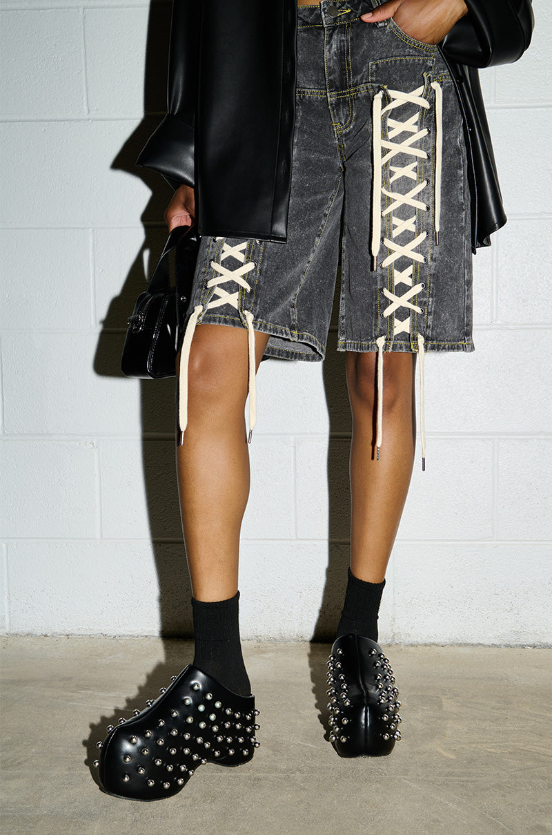 A person is wearing the AZALEA WANG ROMESCO BLACK EMBELLISHED CLOGs adorned with silver spikes, paired with black socks and grey denim shorts featuring white lace-up details on the front. The platform soles add an edgy touch to the ensemble. Dressed in a black faux leather jacket, they pose confidently against a white brick wall while holding a stylish black handbag.
