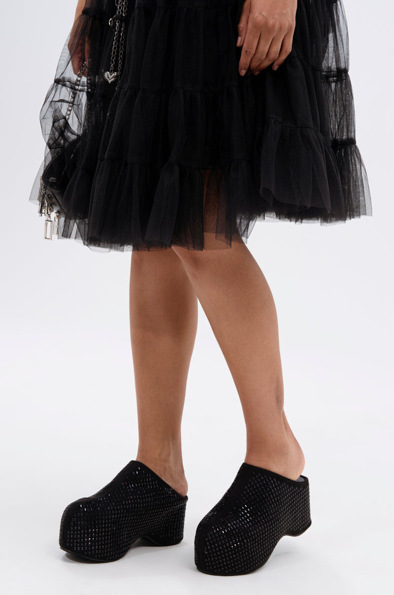 Someone stands against a plain white background wearing a ruffled black tulle skirt and the AZALEA WANG ROMESCO BLING FLATFORM CLOG IN BLACK, which features rhinestone embellishments and a comfort cushion insole.
