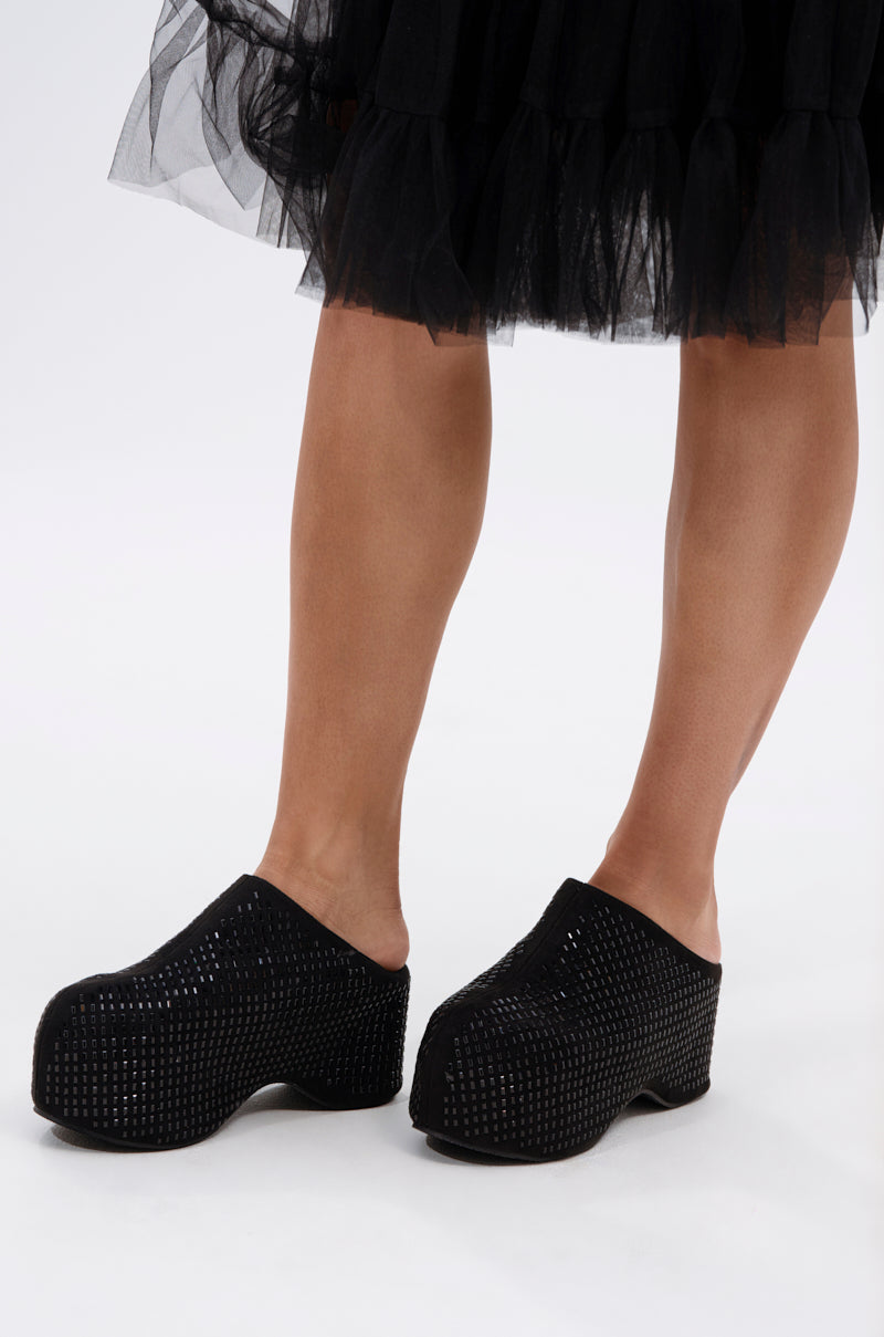 A person stands against a plain white background wearing a black tulle skirt and the AZALEA WANG ROMESCO BLING FLATFORM CLOG IN BLACK, featuring a textured grid pattern and comfort cushion insoles.