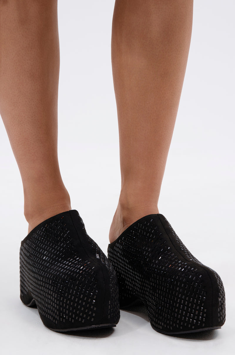 A close-up of a person wearing the AZALEA WANG ROMESCO BLING FLATFORM CLOG IN BLACK, featuring a textured perforated pattern and comfort cushion insole, standing on a white background.