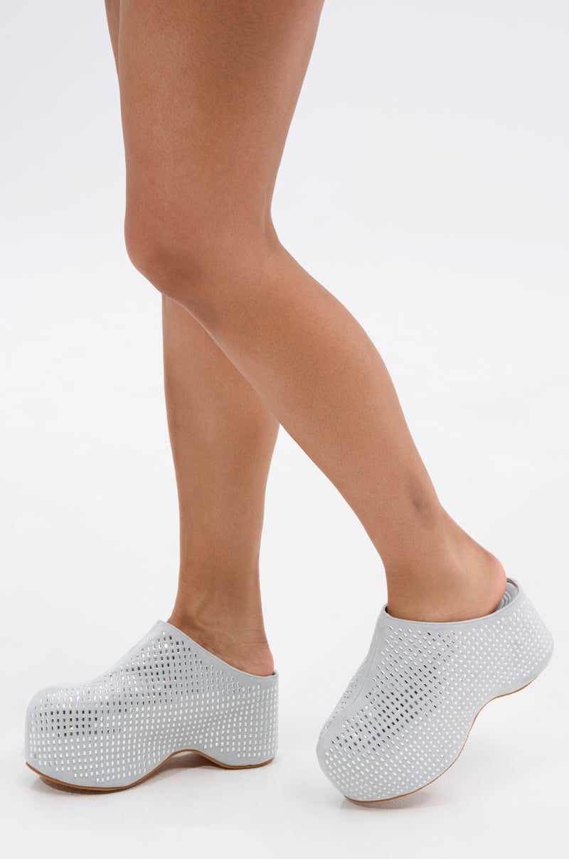 Standing on tiptoe, a person wears the AZALEA WANG ROMESCO BLING FLATFORM CLOG IN GREY—light gray faux suede slip-ons with high, chunky soles; only their legs and feet show against a plain white background.