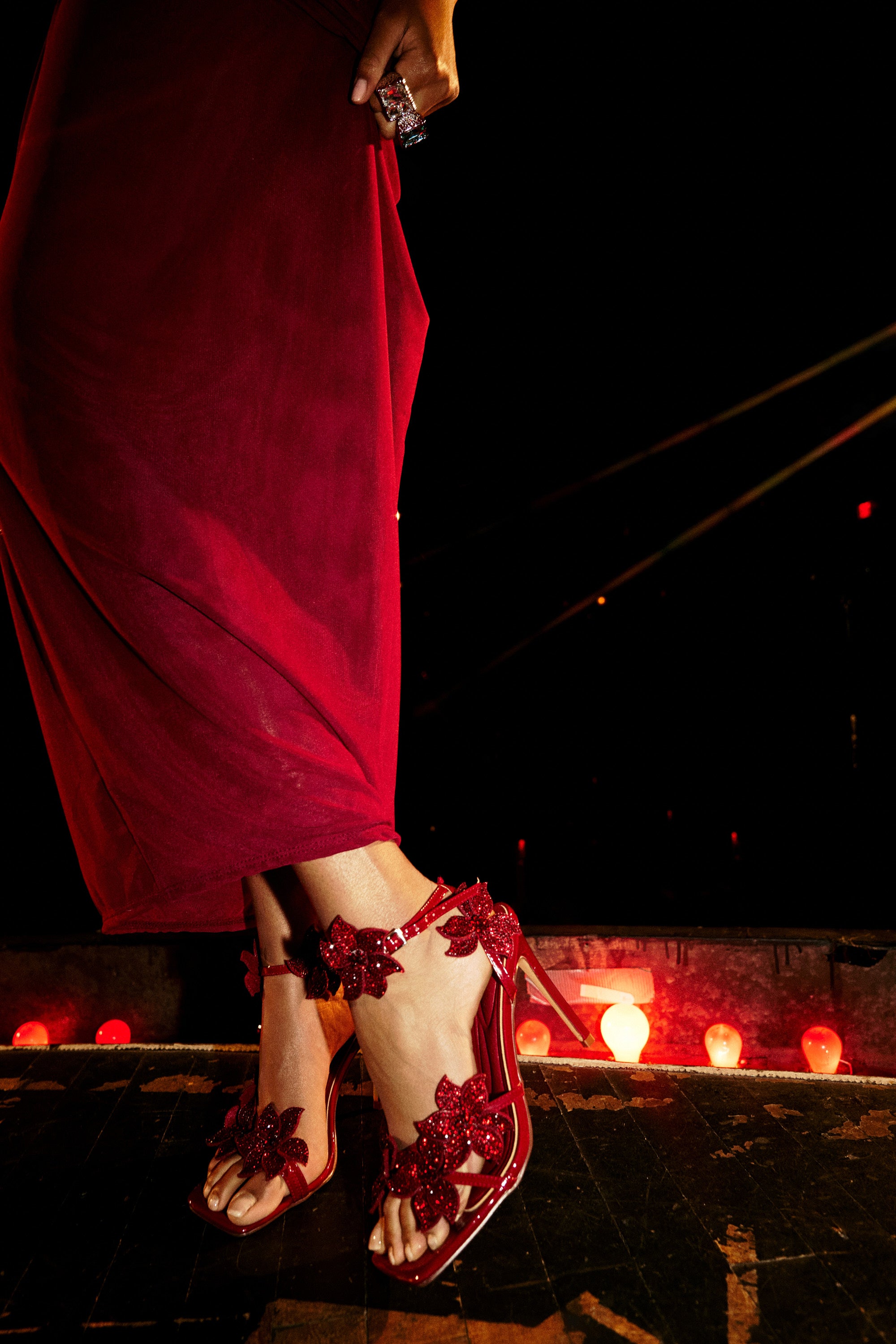 On a dimly lit stage with red lights, a person in a long red dress wears the AZALEA WANG ROMI DARK CHERRY RHINESTONE FLORAL EMBELLISHED SANDAL, whose sparkling flowers add an extra touch of glamour to the scene.