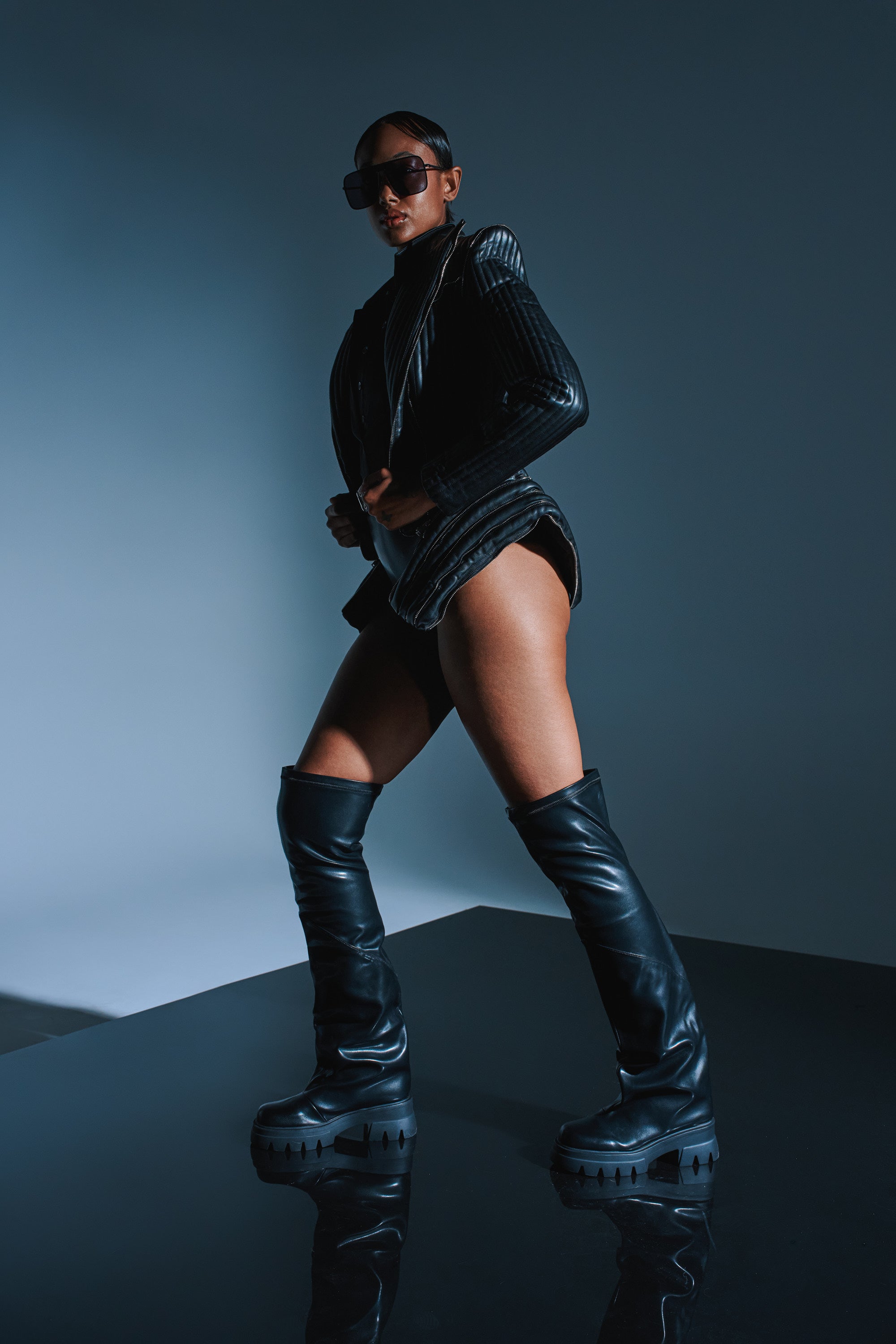 A person poses confidently in the AZALEA WANG RONAN BLACK SLOUCH BOOT, a black jacket, and sunglasses against a dark, minimalist background. Dramatic lighting highlights the bold, modern fashion style.