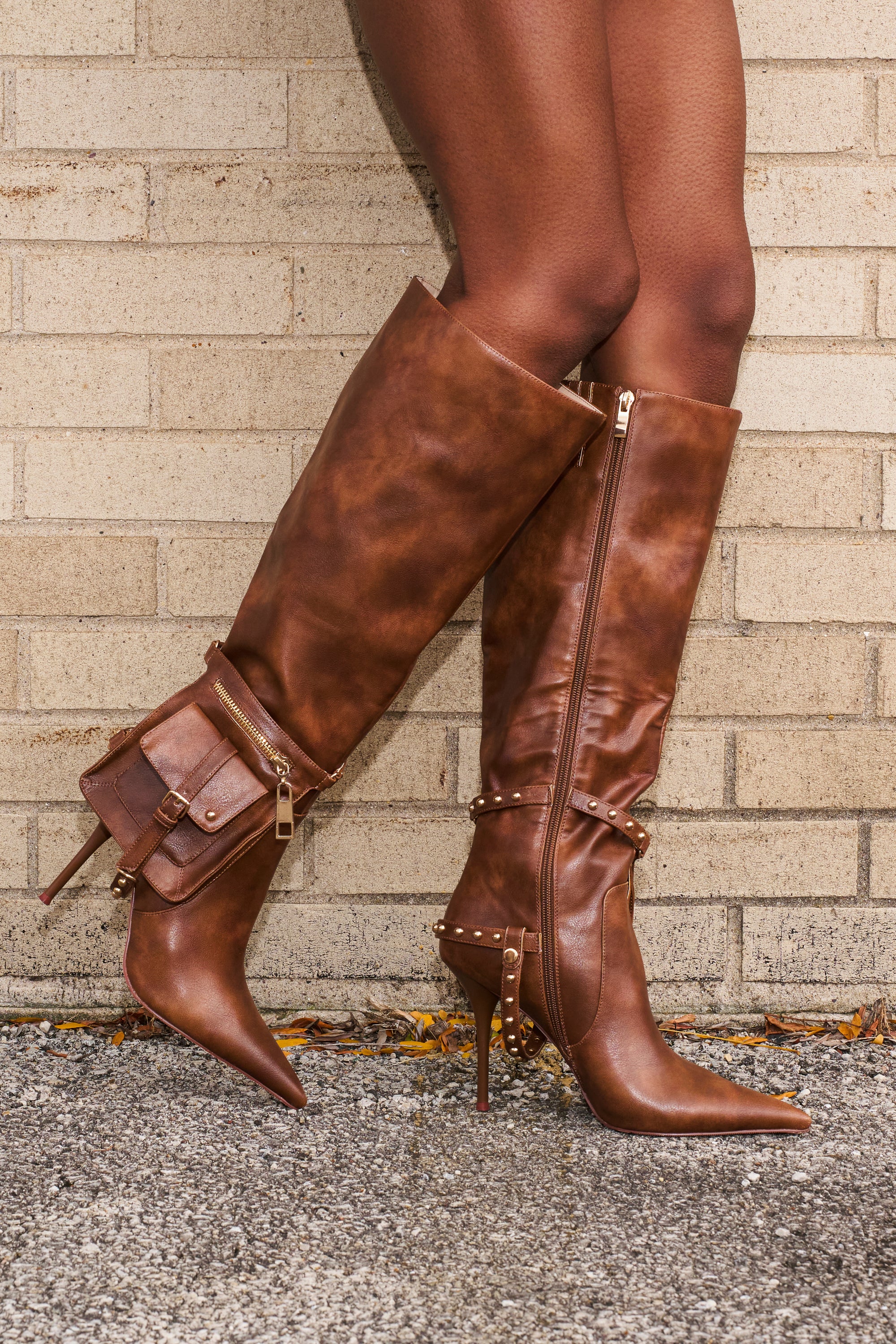 A person in AZALEA WANG RONDEL BROWN DISTRESSED STILETTO BOOTS with gold studs and a matching pouch stands against a light brick wall on gravel.