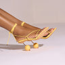 The model wears the RONNIE YELLOW STILETTO STRAPPY SANDALS WITH BALL DETAIL on her feet. These stunning faux leather sandals, featuring crisscross straps and unique, spherical yellow heel bases, stand out against the plain, neutral background.