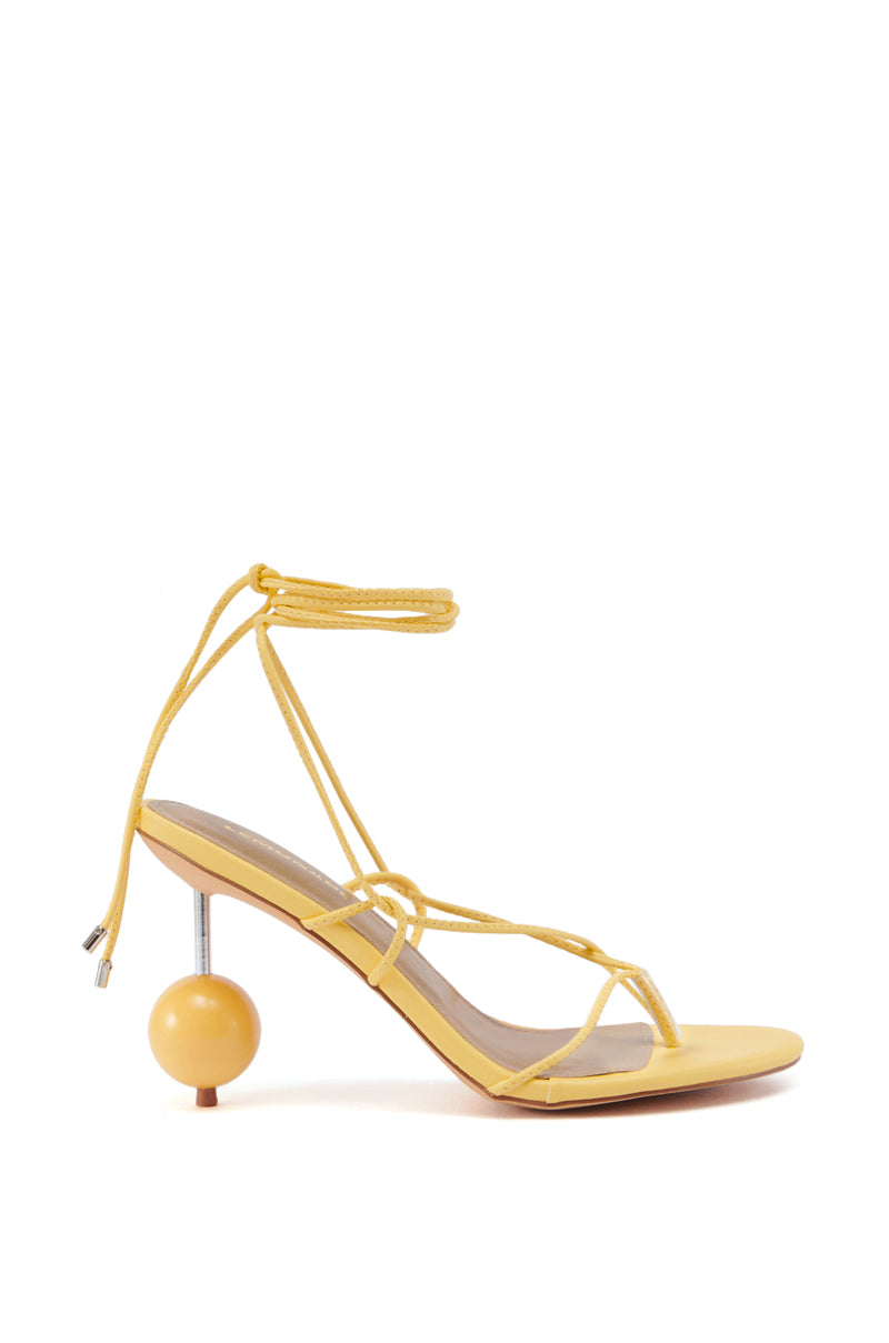 The RONNIE YELLOW STILETTO STRAPPY SANDAL WITH BALL DETAIL is a faux leather sandal designed with a striking heel that features a silver metallic vertical shaft and a yellow spherical base. It includes thin straps that wrap around the ankle in an elegant lace-up style, with both the footbed and sole also in yellow.