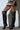 A person stands against a white wall wearing the RONNIE OVER THE KNEE BOOT IN BLACK with a short skirt. The vegan leather, textured thigh-high boots showcase a woven pattern, pointed toes, and stiletto heels, highlighting their elegance alongside the legs.