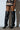 A person stands against a white wall wearing a beige and black skirt with brown accents and the Ronnie Over The Knee Boots in black, vegan leather featuring stiletto heels. Soft lighting casts subtle shadows on the wall.