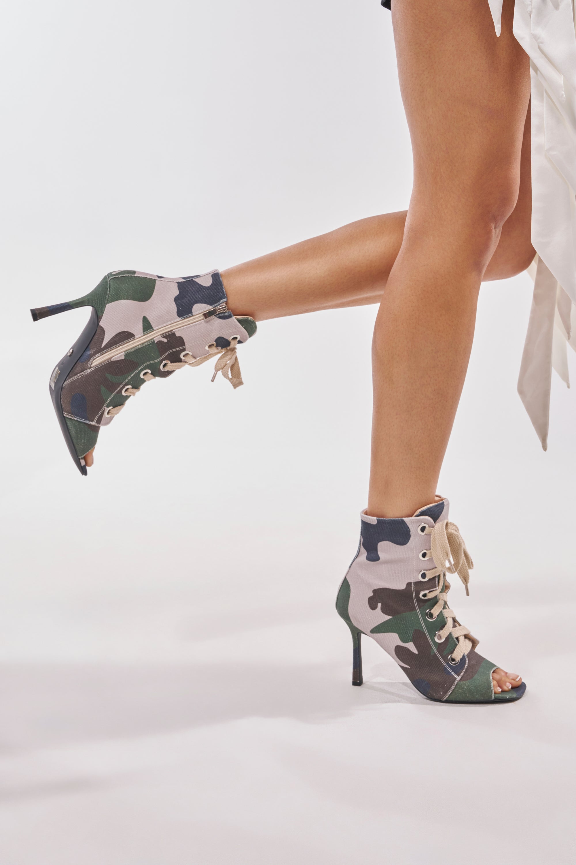 A person models the AZALEA WANG ROOFTOP CAMO OPEN TOE LACE UP BOOTIE—high-heeled, lace-up, with a green, tan, and black camo print—standing on one leg against a light, plain background. 2